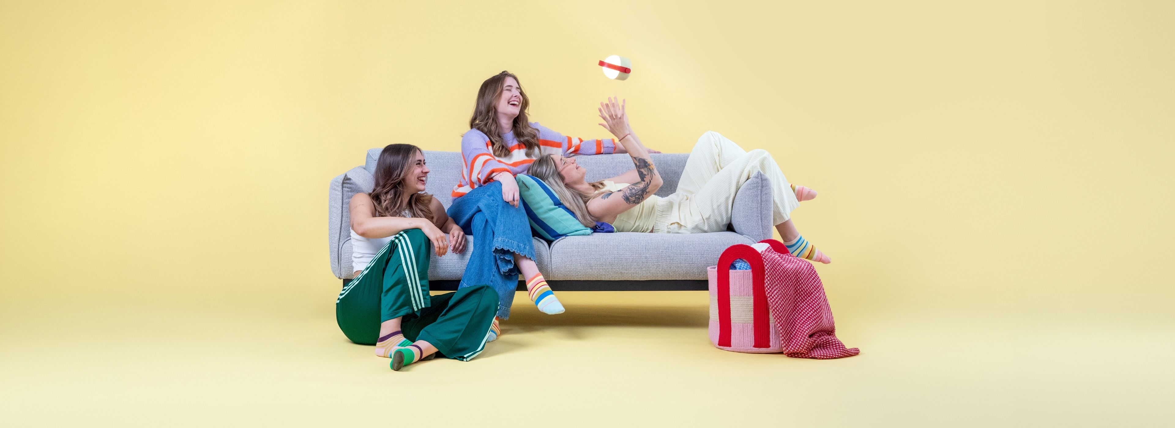 Drei junge Frauen lachen und spielen auf und um ein graues Sofa vor gelbem Hintergrund. Eine liegt entspannt darauf und streckt die Hände nach einer schwebenden, leuchtenden Lampe aus. Zwei Freundinnen sitzen daneben.