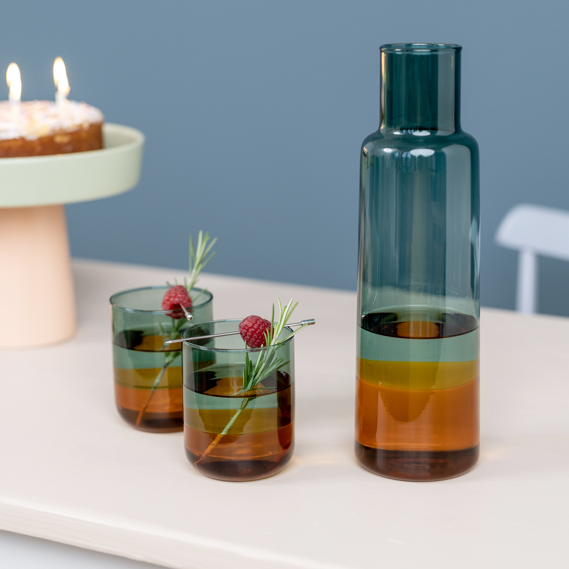 Two 'amber' glasses with drink, decorated with raspberry and rosemary, next to a matching carafe on a table. A cake with candles in the background.