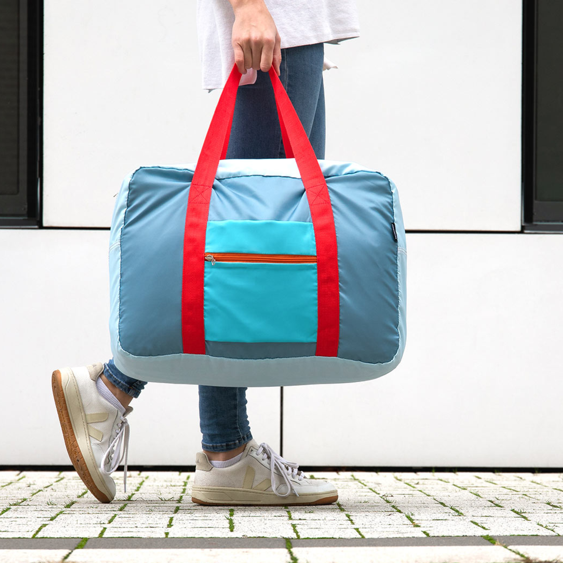 Una persona trasporta la borsa da viaggio "Tom" di colore azzurro con cinghie rosse. La borsa ha una tasca esterna blu con cerniera. Lo sfondo è una parete chiara.