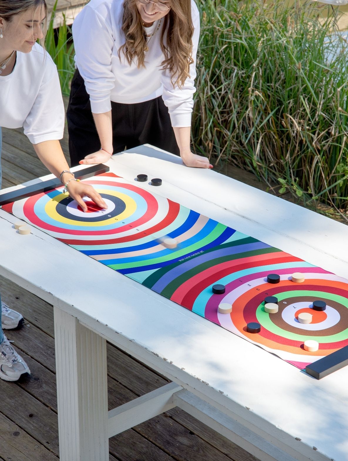 Zwei Frauen spielen ein Wurfspiel auf einem weißen Tisch. Eine schiebt einen Stein auf eine bunte Zielmatte mit Ringen, weitere Steine liegen dort. Im Hintergrund grüne Wasserpflanzen.