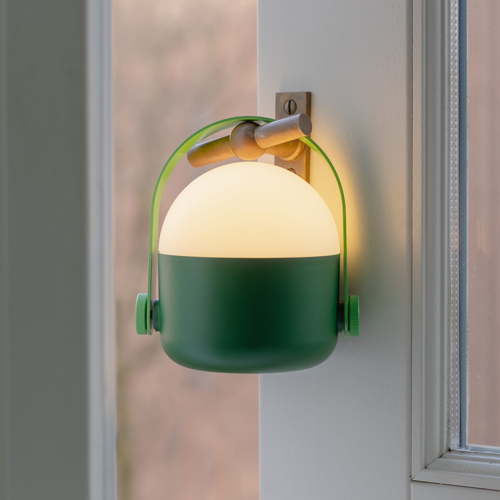 Una lámpara verde con asa y luz blanca cálida está montada en una pared junto a una ventana.