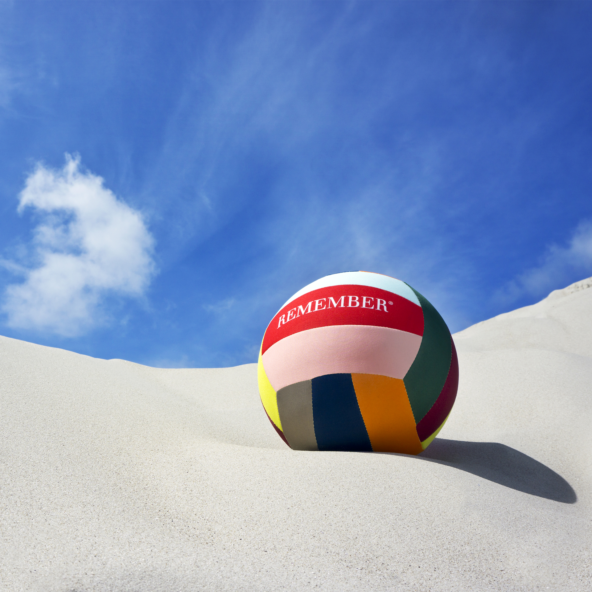 A colourful leisure ball with the inscription "REMEMBER" lies in the sand under a blue sky with clouds.