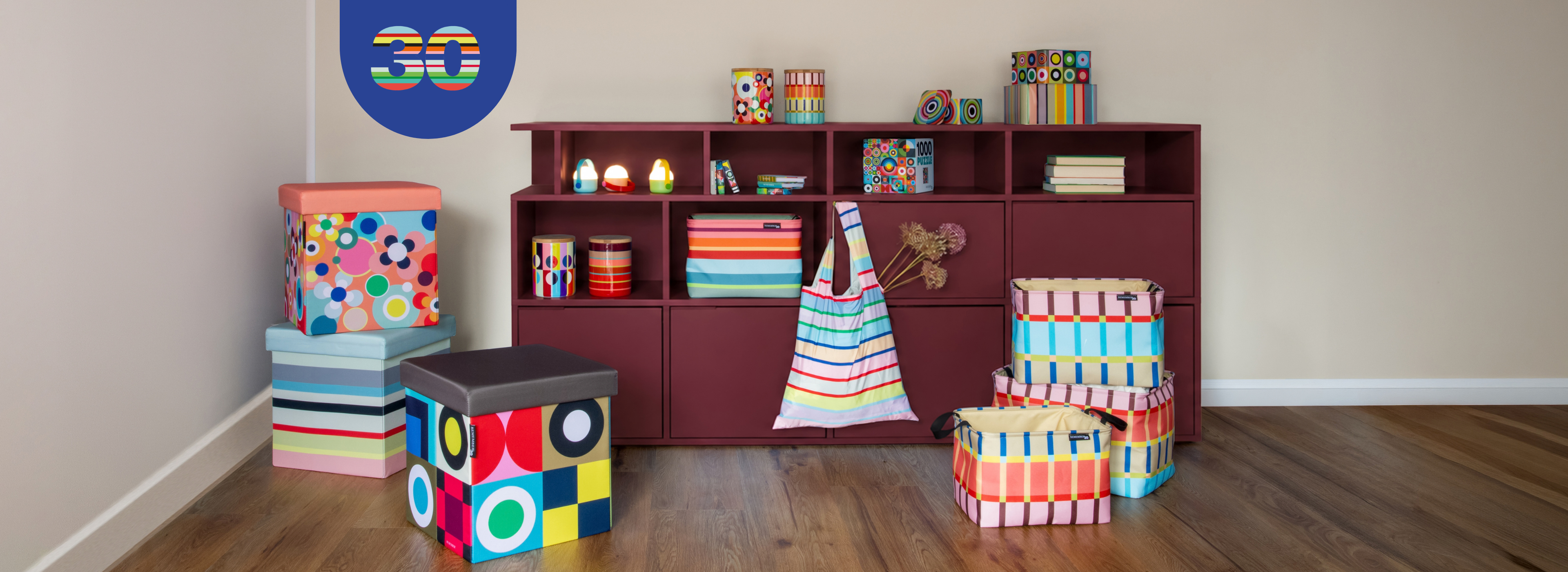 Divisão com estante vermelho-escura e acessórios coloridos: latas às riscas e com padrões geométricos, livros, puzzle, bancos e caixas florais, às riscas e em cubos. Sacola pendurada. Logótipo 30 anos na parede.