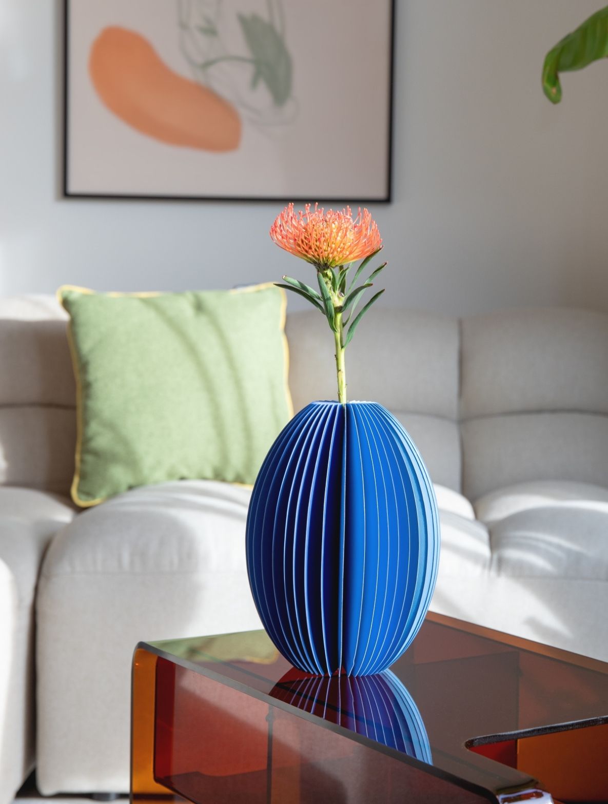 Eine orangefarbene Protea mit feinen Blütenblättern steckt in einer geriffelten blauen Ovalvase. Die Vase steht auf einem transparenten Couchtisch mit roten Kanten. Im Hintergrund: helles Sofa, grünes Kissen, abstraktes Wandbild.
