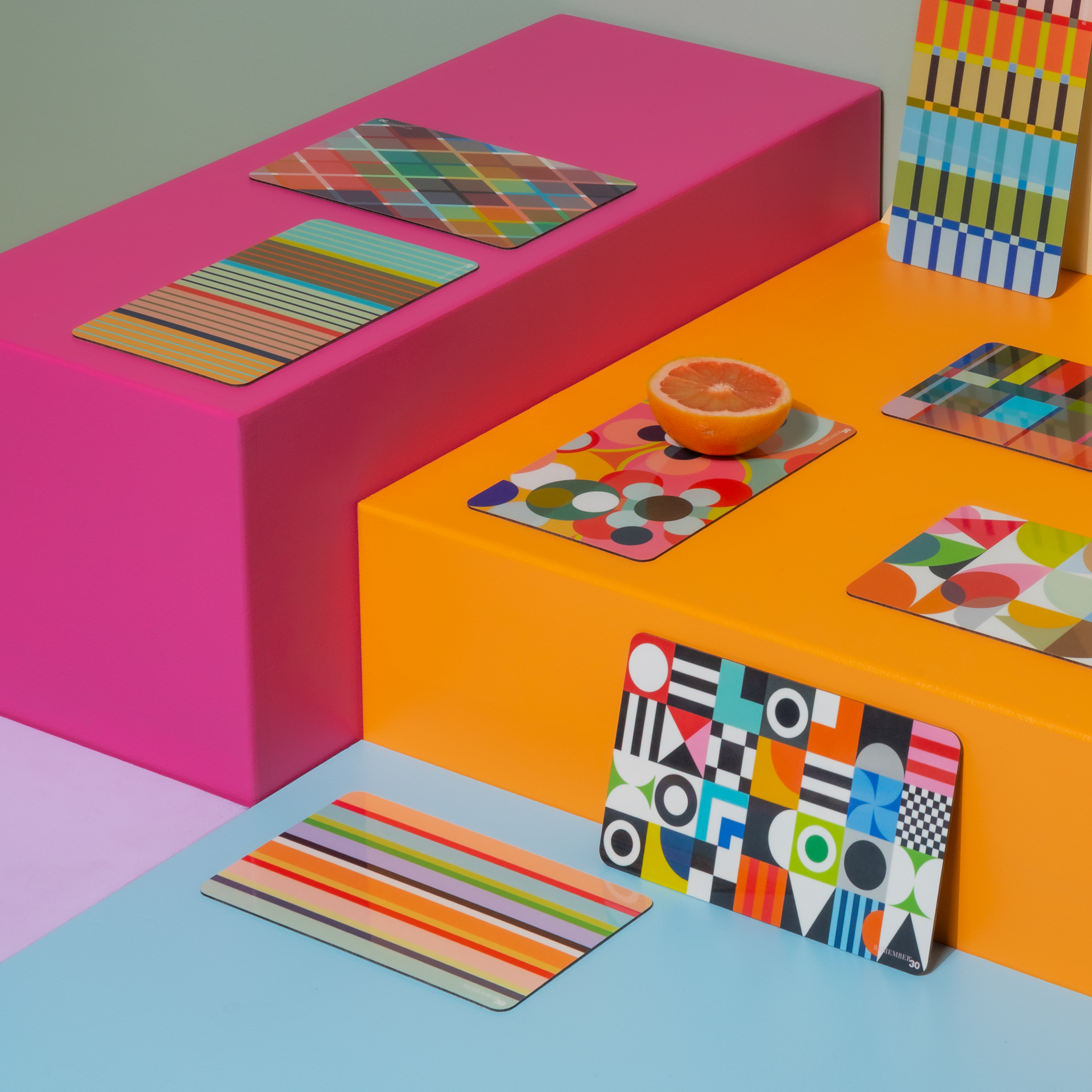 Mehrfarbiges Arrangement: Geometrische Muster, Streifen und Punkte auf Platzsets, platziert auf pinkfarbenen, orangen und blauen Plattformen. Eine halbe Orange ergänzt das Ensemble.