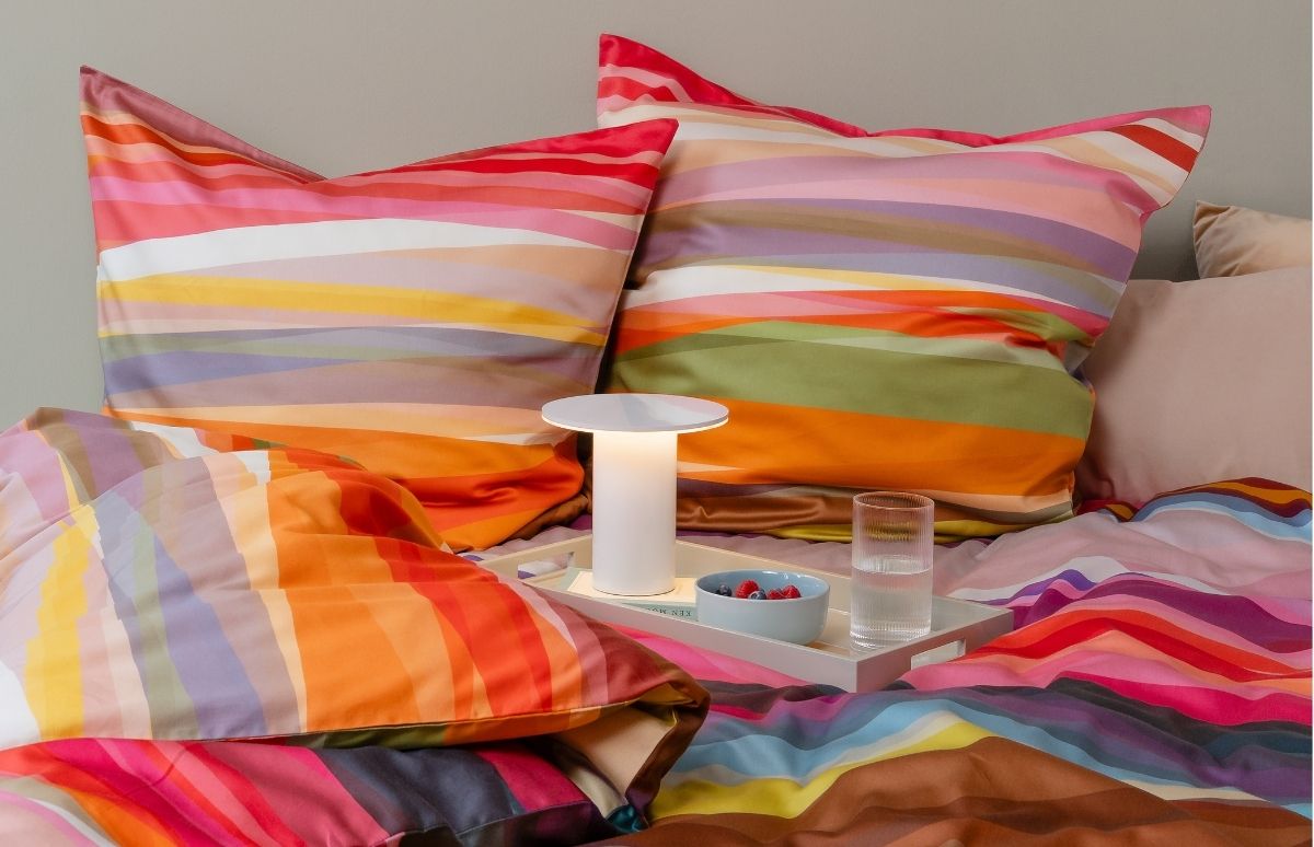 Ein gemütlich gemachtes Bett mit horizontal gestreifter Bettwäsche in lebhaften Farben wie Orange, Pink, Gelb und Grün. Auf der Decke steht ein helles Tablett mit beleuchteter Tischlampe, blauer Beerenschale und Glas Wasser.