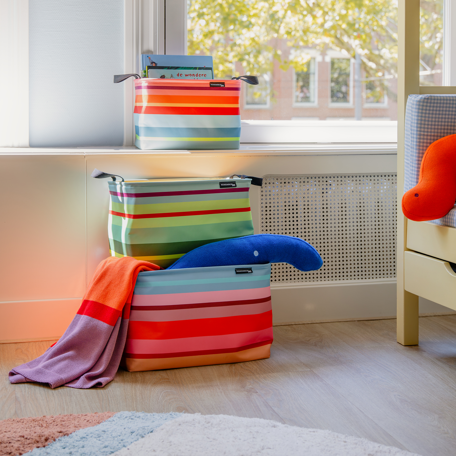 Drei gestreifte „Nesa Remember“-Aufbewahrungskörbe in drei Größen in einem hellen Raum: klein mit Büchern auf der Fensterbank, zwei größere am Boden mit Spielzeug/Decke; bunte Streifen, Griffe.