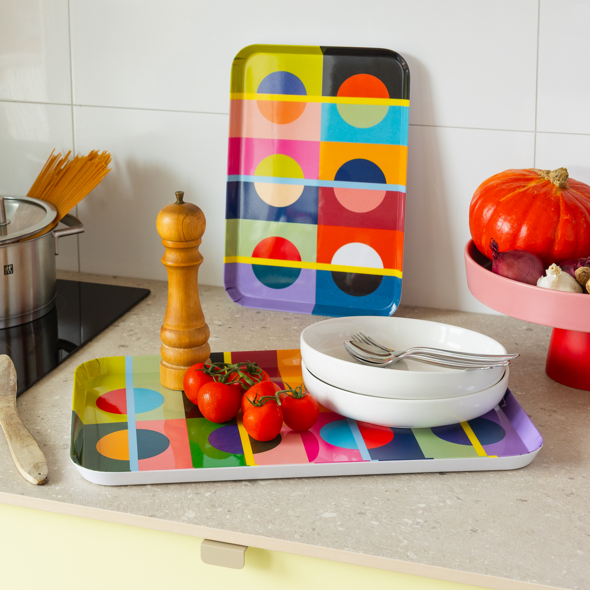 Due vassoi rettangolari con un vivace motivo geometrico Orb composto da quadrati e semicerchi nei colori rosa, blu e arancione in una cucina moderna. Uno è appoggiato con pomodorini e ciotole bianche, l’altro è appoggiato al rivestimento in piastrelle.