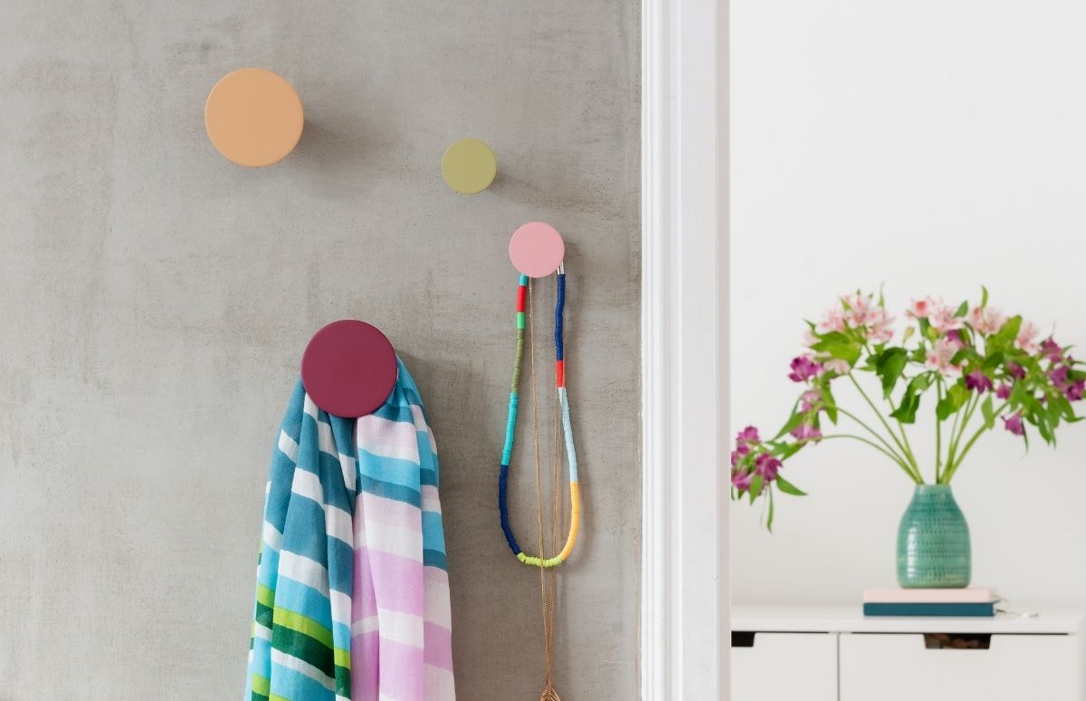 Drei bunte, runde Wandhaken an grauer Betonwand. Ein weinroter Haken hält einen gestreiften Schal in Blau, Weiß, Pink und Grün. Ein hellpinker Haken trägt zwei Ketten. Rechts: Türzarge und Kommode mit Vase voller pinker und lila Blumen.