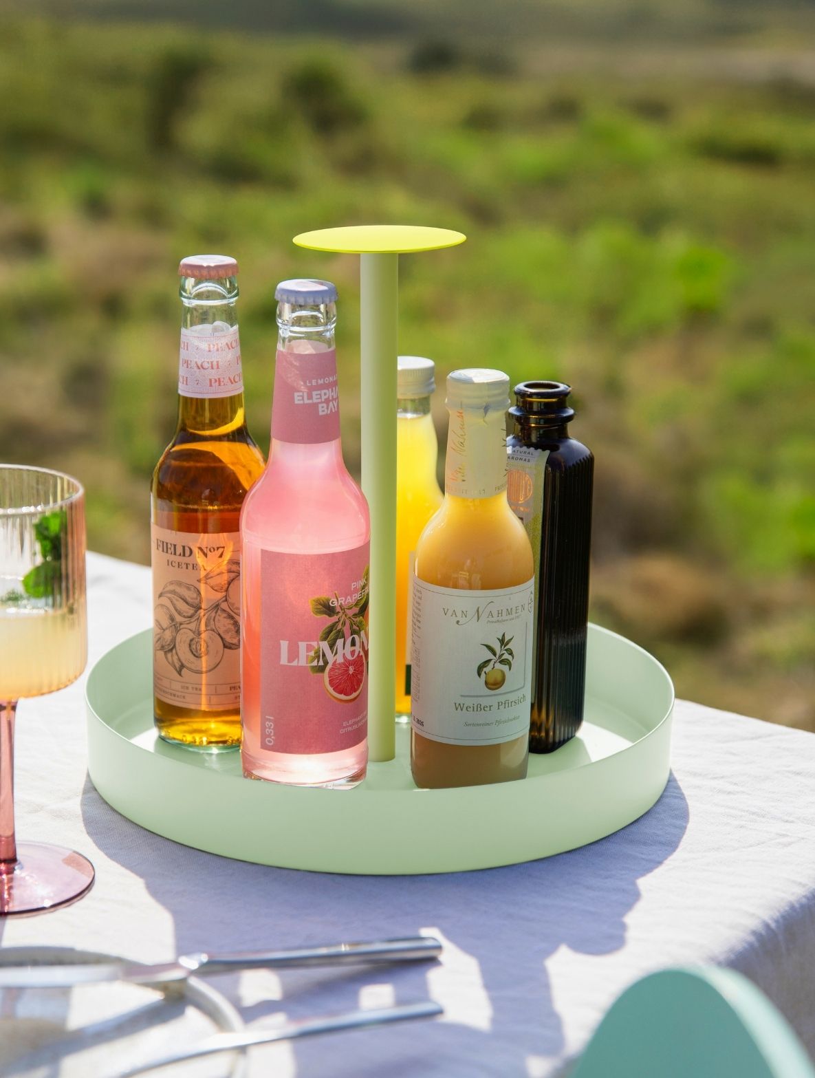Hellgrünes Serviertablett auf weißem Tisch im Freien mit Getränkeflaschen: Eistee, rosa Grapefruitlimonade, weißer Pfirsichsaft. Mittig limonengrüner Ständer mit gelber Ablage. Glas mit Minze und Besteck vorn, hinten grünes Bokeh.