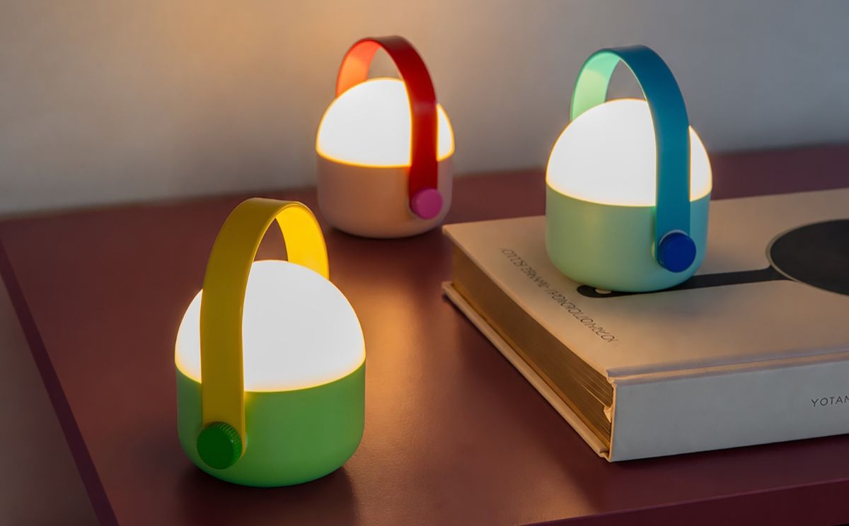 Três lanternas modernas e luminosas sobre mesa avermelhada. À frente: base verde, pega amarela, botão giratório verde. Atrás: base rosa-claro, pega vermelha. À direita, ao fundo: base azul-clara, pega azul. Ocultas pelo livro.