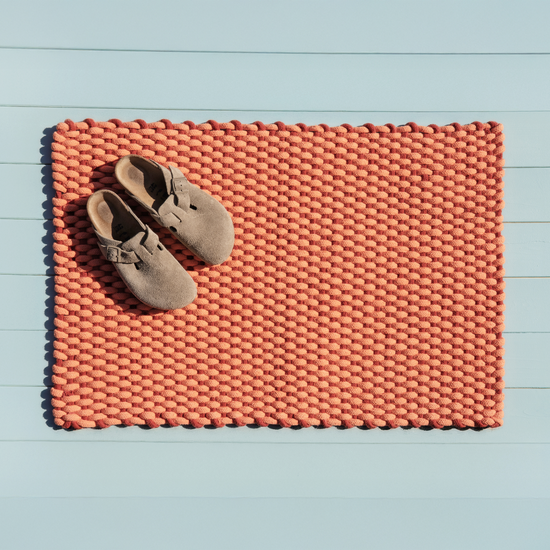 Alfombra tejida a mano en color coral con relieve sobre fondo azul. Hay un par de zapatos en el lado izquierdo de la alfombra.