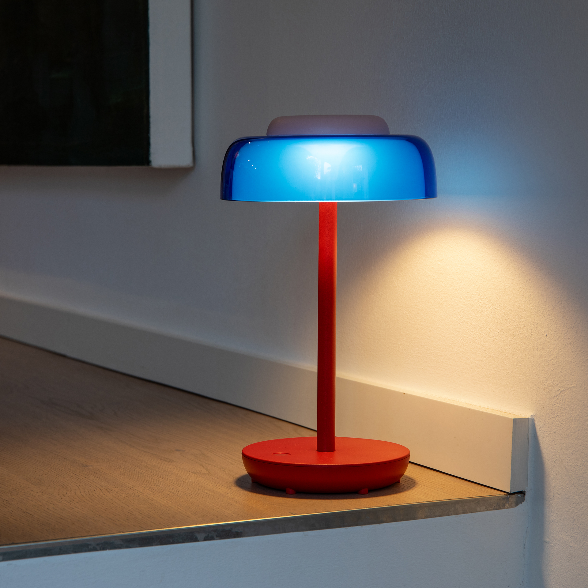 Een draadloze tafellamp genaamd Juno, met een slanke rode metalen voet en stang. De doorzichtige, blauwe kunststof kap omhult een roze, rond aanraakdimmvlak. Op de rode voet is een subtiel aan/uit-symbool te zien.