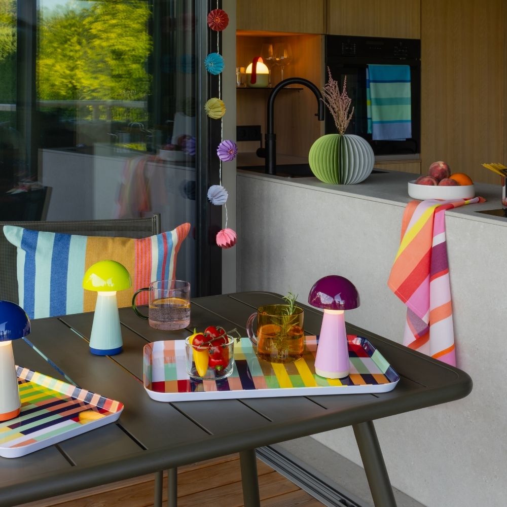 Gedeckter Balkontisch mit drei leuchtenden Pilzlampen in Blau, Hellgrün und Lila. Zwei bunte Tabletts mit Getränken und Snacks. Im Hintergrund moderne Küche mit Spülbecken und grüner Faltvase, dahinter üppige Bäume.