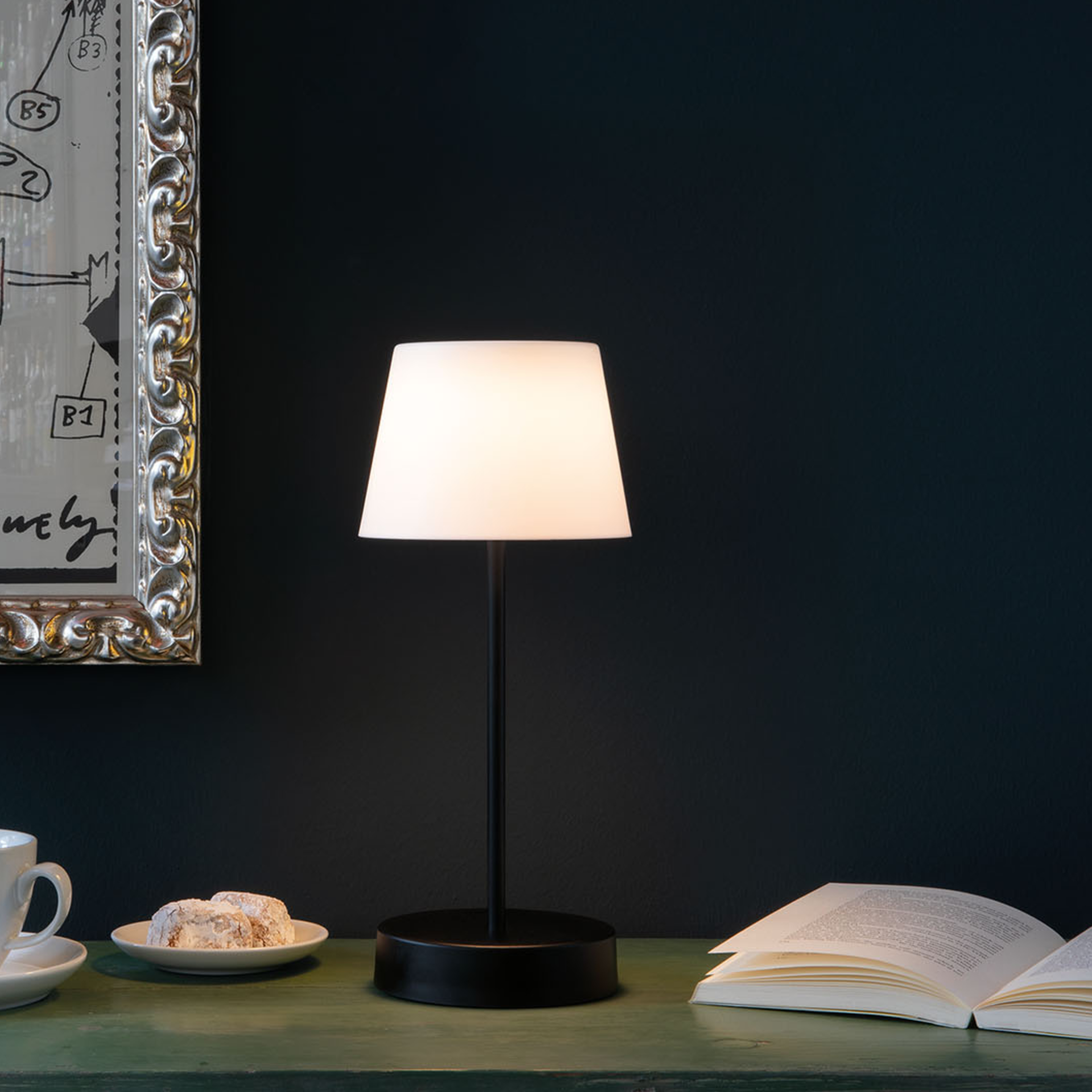 Table lamp Oscar 'pure' on a table next to an open book and a cup. The lamp has a black base and a white, luminous cap.