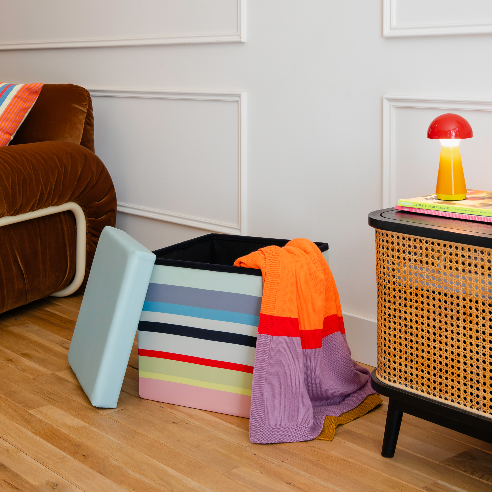 Bunte gestreifte Sitzbox auf Holzboden, hellblauer Deckel daneben. Aus der offenen Box ragt eine leuchtend orange Decke mit lila und roten Akzenten. Im Hintergrund brauner Samtsessel, Beistelltisch mit roter Pilzlampe.
