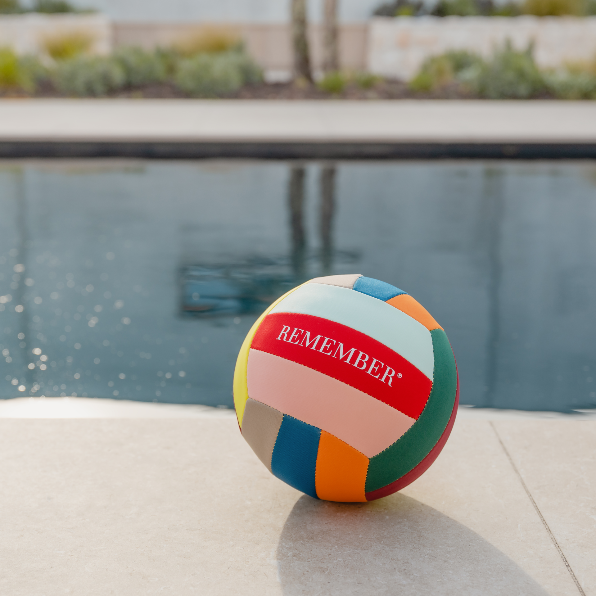A colourful leisure ball with the lettering "REMEMBER" lies on the edge of a pool. Plants can be seen in the background.