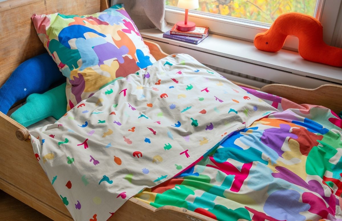 Kinderbett mit bunter Decke und Kissen; oben blaues und grünes Stofftier. Fensterbrett: rote Lampe, Bücher, Kuscheltier. Draußen unscharfe Herbstbäume.