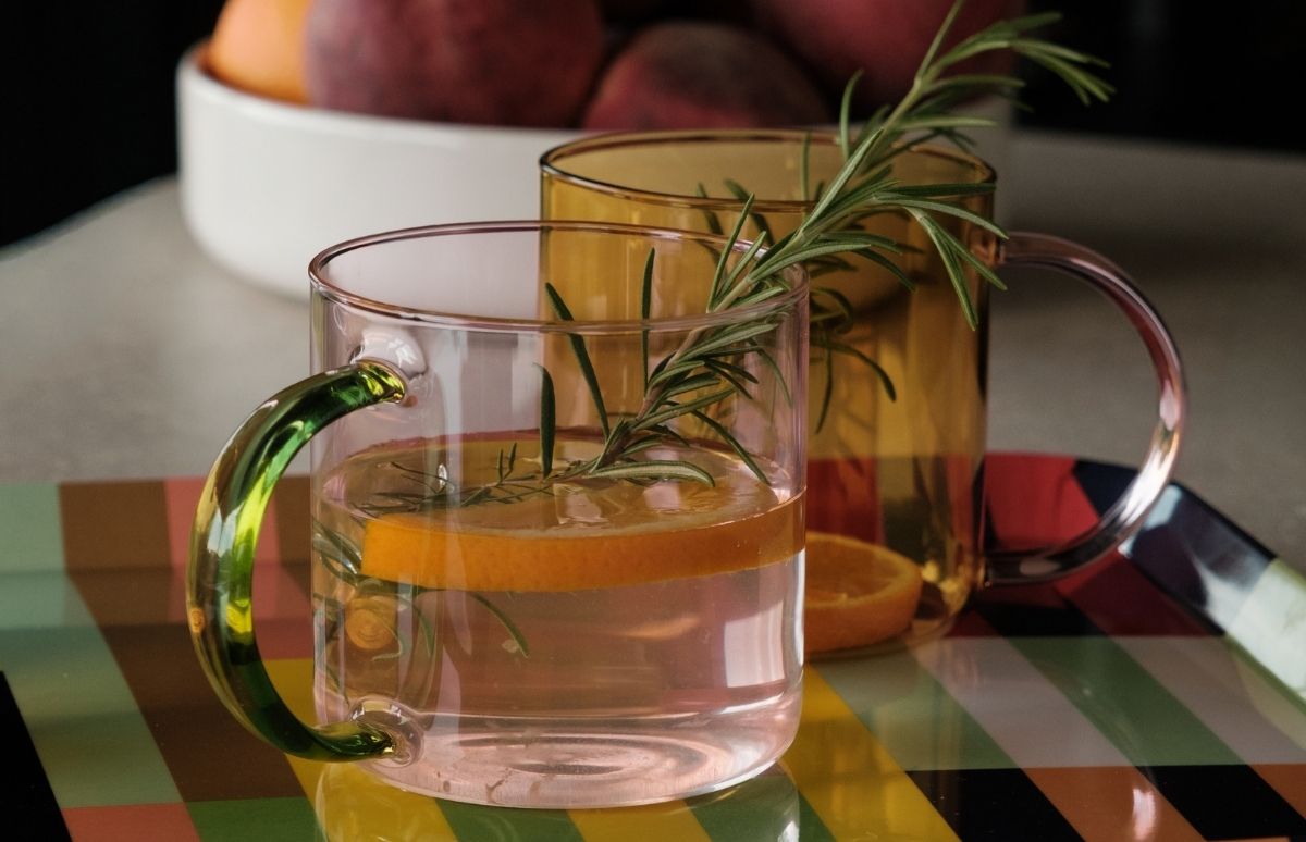 Zwei farbige Glastassen auf gestreiftem Tablett. Vorn eine blassrosa Tasse mit grünem Henkel, Wasser mit Orangenscheibe und Rosmarinzweig. Dahinter eine bernsteinfarbene Tasse mit klarem Henkel und Rosmarin. Im Hintergrund Früchte in weißer Schale.