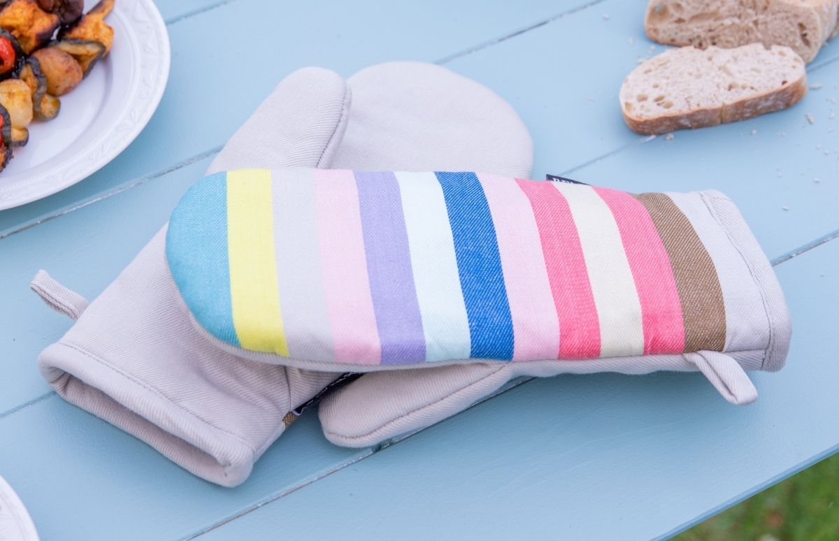 Ein Paar hellbeige Ofenhandschuhe liegt auf einem hellblauen Holztisch. Einer zeigt auf der Handfläche ein Streifenmuster in Pastelltönen wie Blau, Gelb, Rosa und Lila. Im Hintergrund: gegrilltes Gemüse und Brotscheiben.