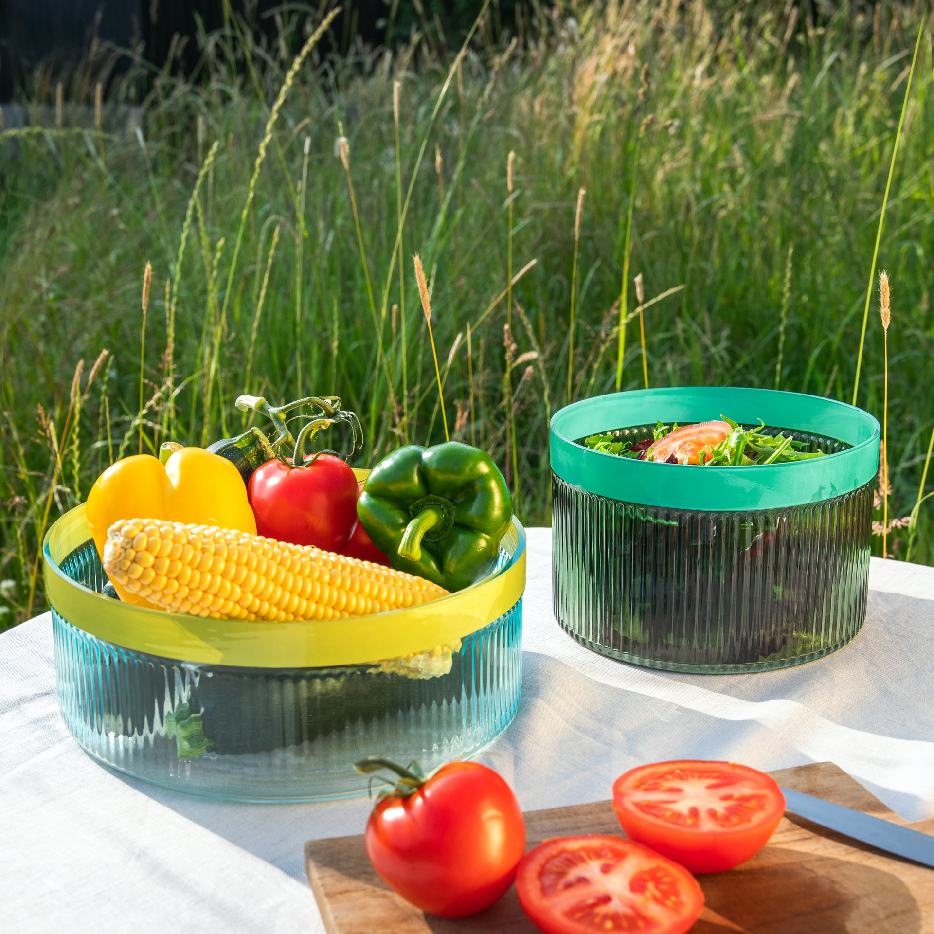 Twee glazen schalen met groeven, een groene en een blauwe, gevuld met salade en groenten, op een buitentafel.