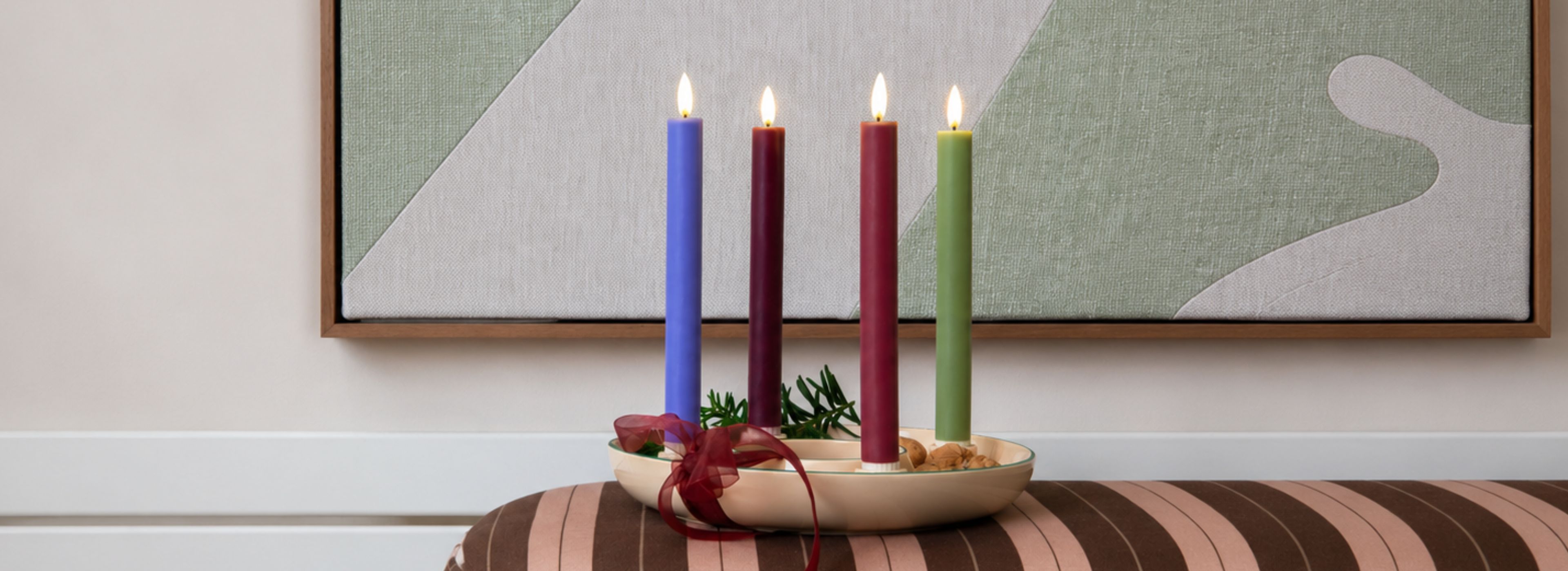 Vier brennende Kerzen (blau, zwei dunkelrot, grün) in einem cremefarbenen Adventsgeschirr. Verziert mit Tannengrün, Naturmaterialien und weinrotem Band. Auf gestreiftem Kissen; abstraktes Bild in Hellgrün und Beige.