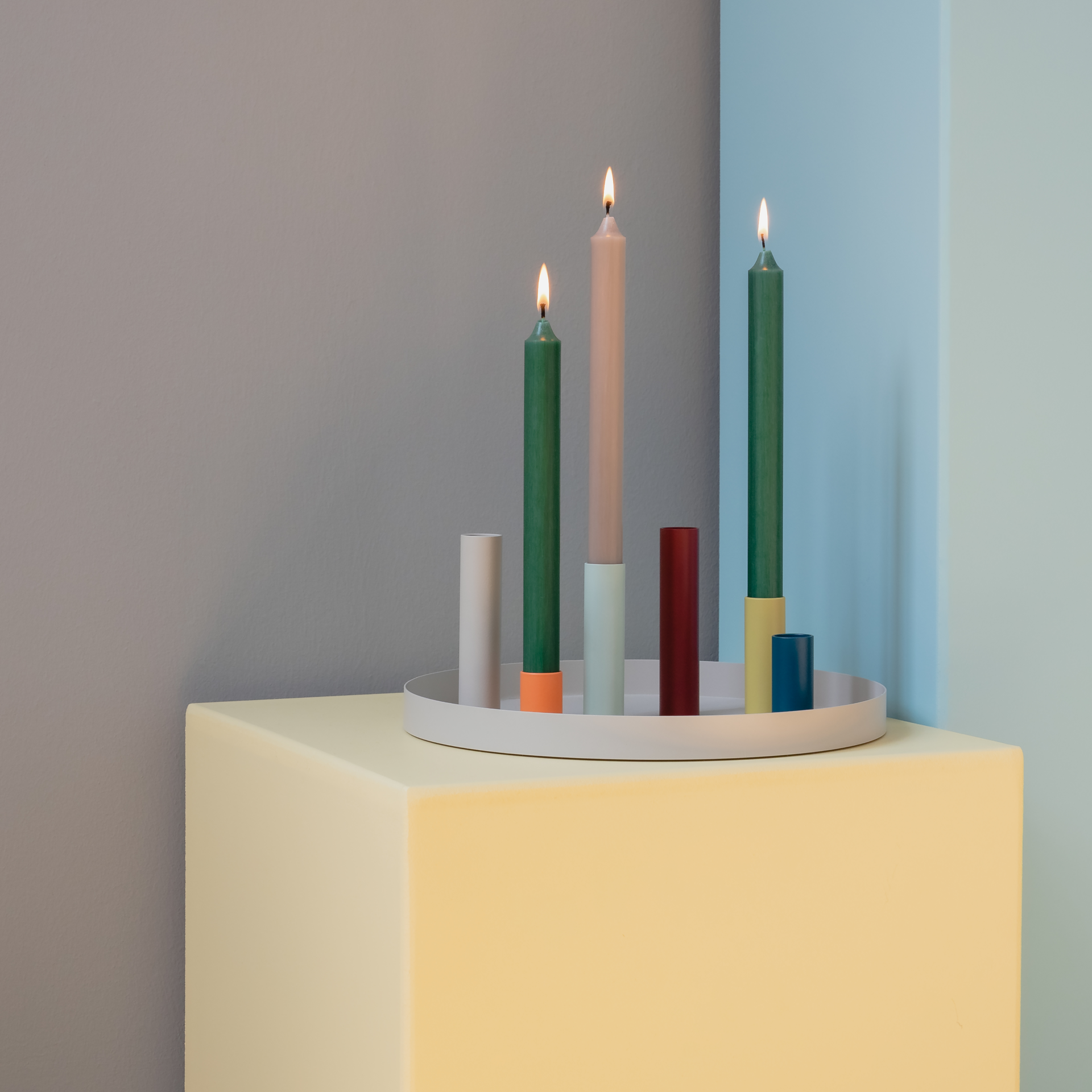 Sei portacandele colorati con tre candele accese su un vassoio rotondo, presentati su un tavolo giallo di fronte a pareti colorate.