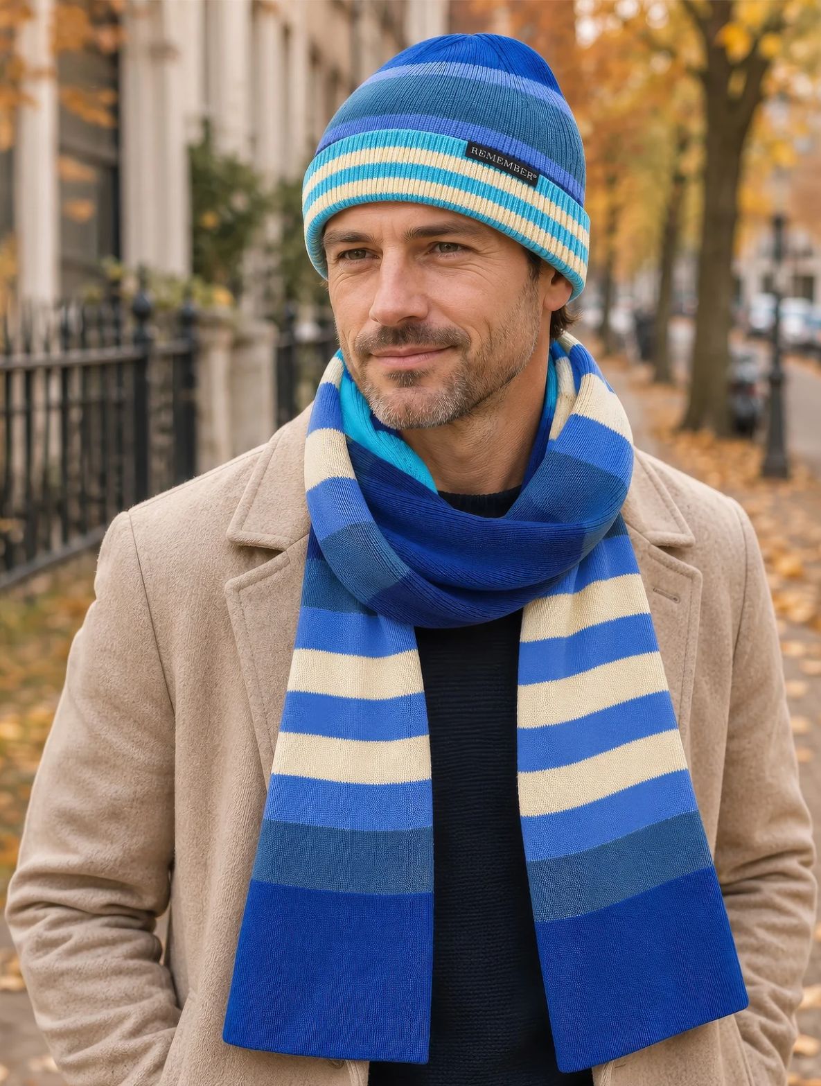 Ein Mann mit gestreiftem blau-cremefarbenem Beanie und passendem Schal geht auf einer herbstlichen Straße. Er trägt einen beigen Mantel, blickt leicht nach rechts und lächelt dezent. Im Hintergrund Bäume mit goldenen Blättern.