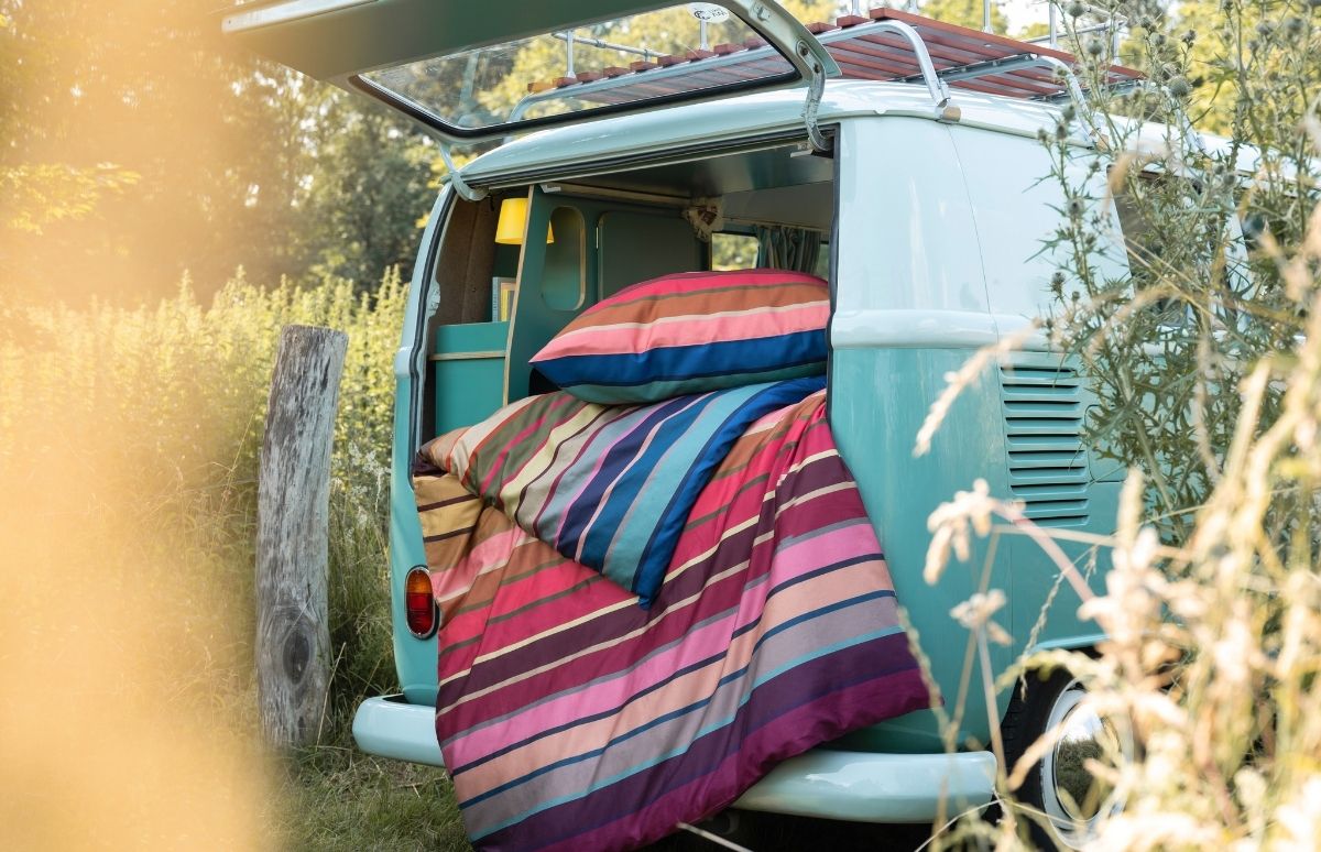 Ein geöffneter, hellblauer Campingbus steht in einem sonnigen, grünen Feld. Im Heck des Busses liegen Decke und Kissen mit bunten Querstreifen in Pink-, Blau- und Erdtönen, bereit für eine Übernachtung.