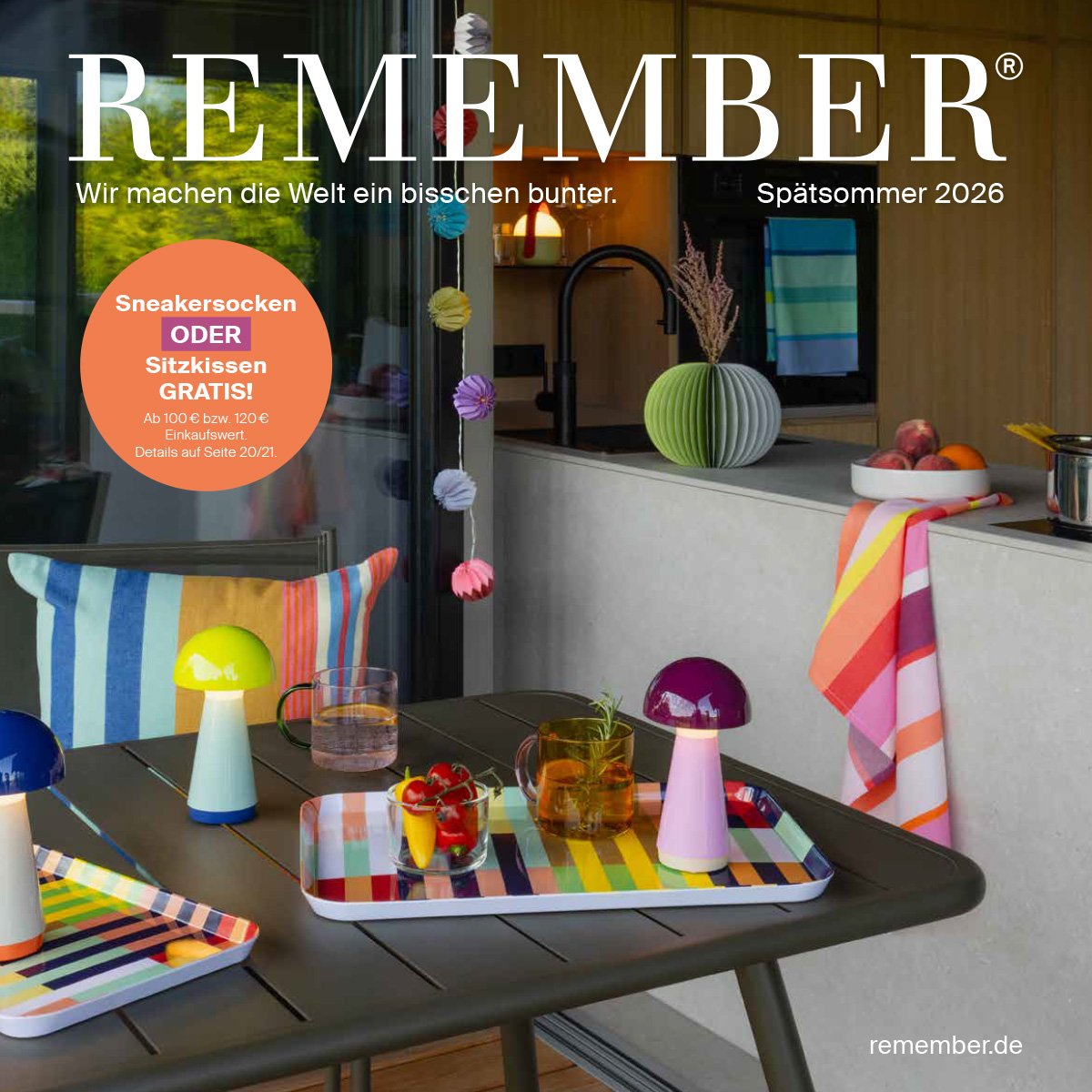 Stilvolle Terrasse mit dunklem Tisch: bunte Pilzleuchten, Getränke und Snacks auf gestreiften Tabletts. Im Hintergrund moderne Küche mit Obstschale, gestreiftem Handtuch und grüner Faltvase. Bunte Girlanden. REMEMBER.