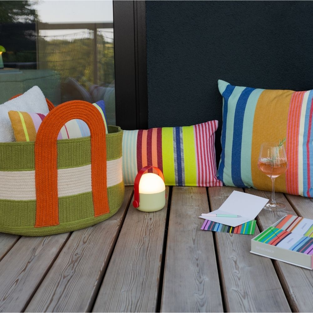 Gemütliche Outdoor-Oase auf einer Holzterrasse mit farbenfrohen gestreiften Kissen in Blau, Orange, Gelb und Grün. Ein olivgrüner Korb mit weißen Streifen und orangefarbenen Henkeln mit Textilien. Lampe, Rosé mit Rosmarin, Notizbuch.