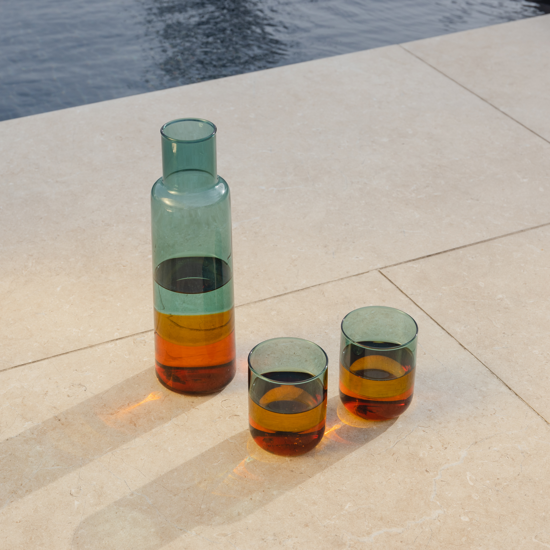 Two 'amber' glasses and a carafe stand on a beige stone surface. The glasses are filled with liquid that shimmers in various shades of amber. Water is visible in the background.