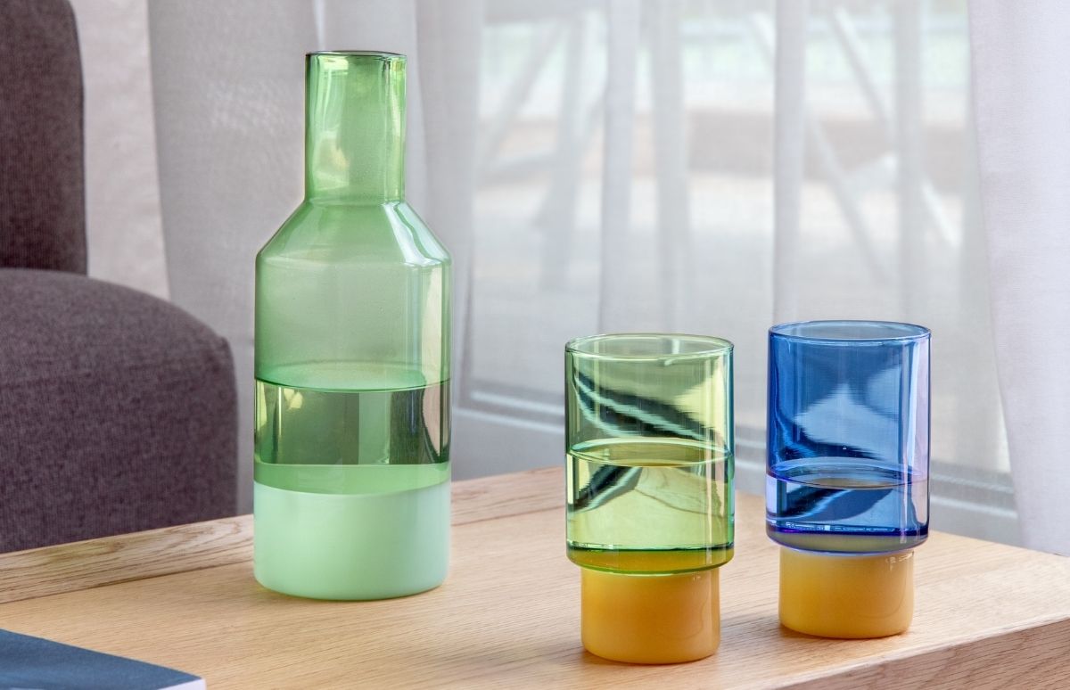 Hohe grüne Glaskaraffe und zwei kleinere bunte Trinkgläser auf hellem Holztisch. Karaffe mit drei Grüntönen von transparent bis matt hellgrün. Gläser oben grün/blau transparent, unten gelb. Mit Wasser gefüllt vor Fenster mit weißen Vorhängen.