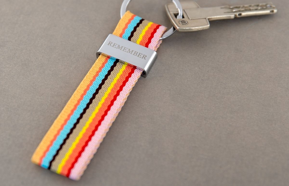 Schlüsselbund mit buntem, gestreiftem Stoffband auf grauer Fläche; silberne Halterung mit „REMEMBER“ und ein Schlüssel am Ring.