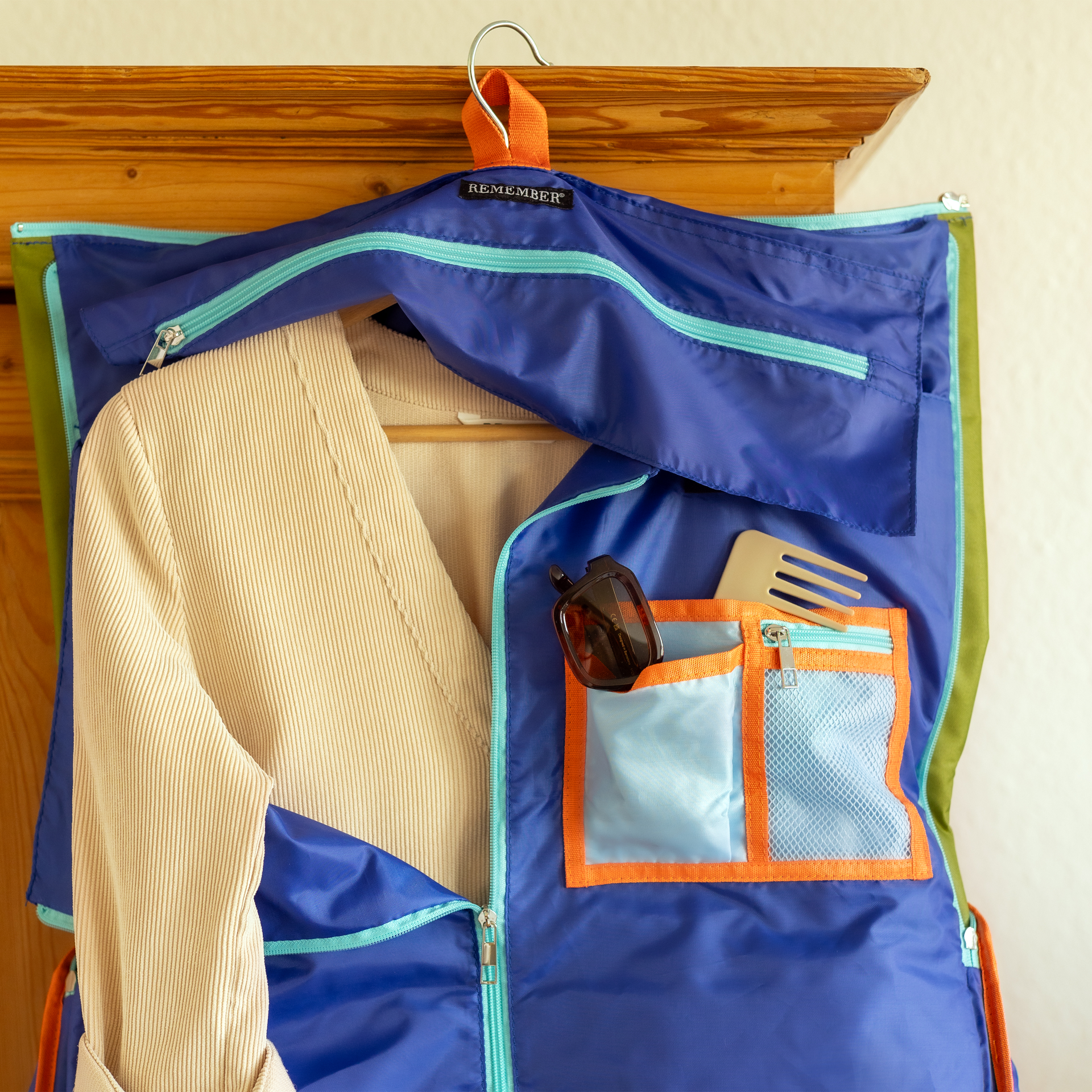 A blue Lennard travel bag, opened out and featuring turquoise zips, is hanging from a wooden clothes hanger. A beige corduroy garment is hanging inside the bag. On the right: organiser compartments containing sunglasses and a comb.