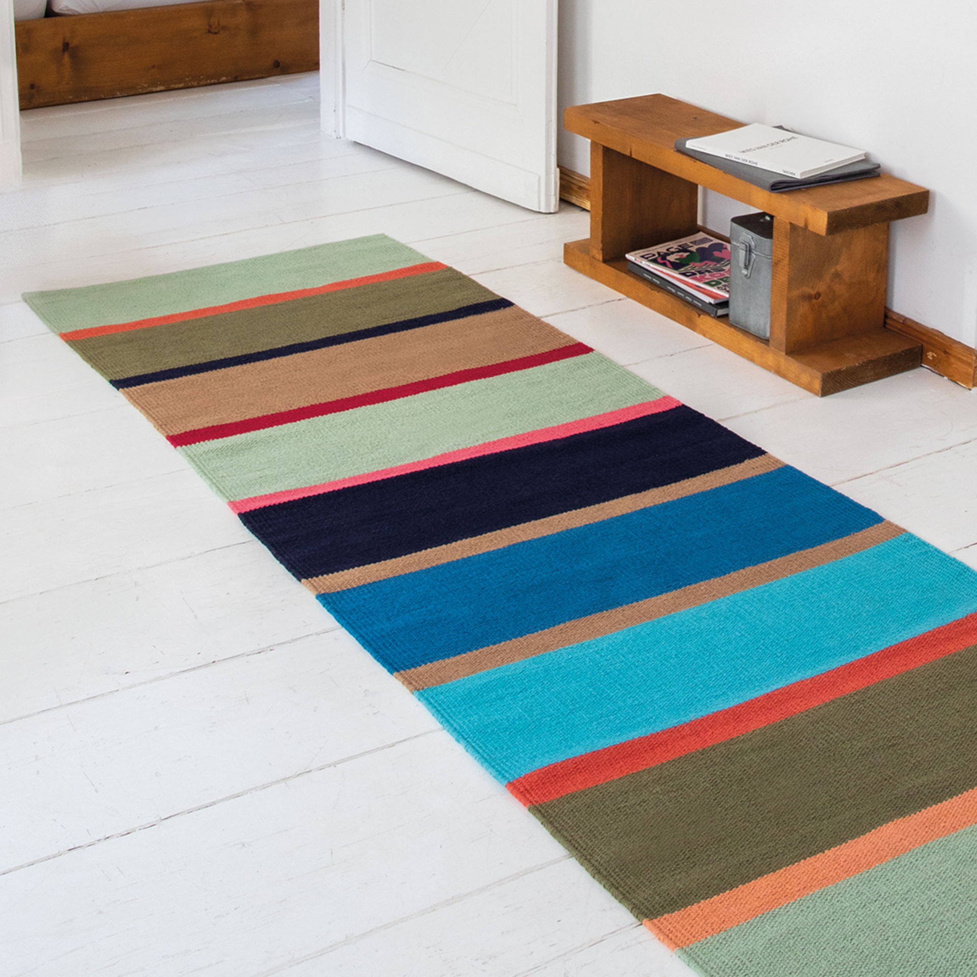 Tapis de sol à rayures longues 'Costa' en coton sur un sol en bois blanc. Couleurs : blanc, rouge et bleu : Vert, brun, bleu, rouge. En arrière-plan, une étagère en bois et une porte blanche ouverte.