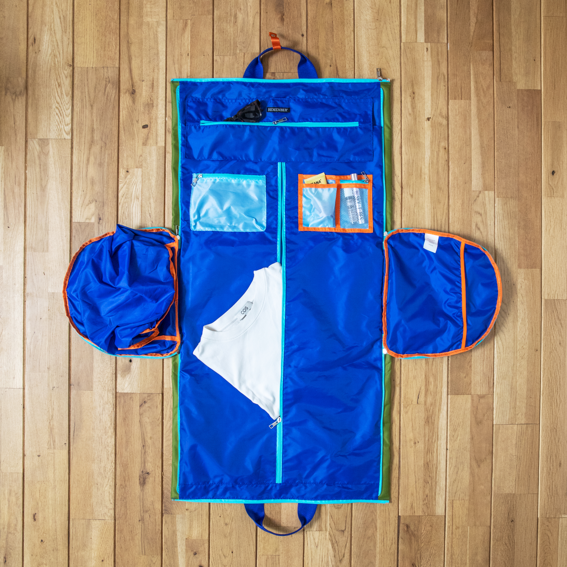 Große blaue Reisetasche Lennard, geöffnet auf hellem Holzboden. Im geräumigen Inneren: weißes T-Shirt, Sonnenbrille und Toilettenartikel in Fächern. Runde Seitenfächer, wohl für Schuhe, ausgeklappt – Packungssystem für Reiseutensilien.