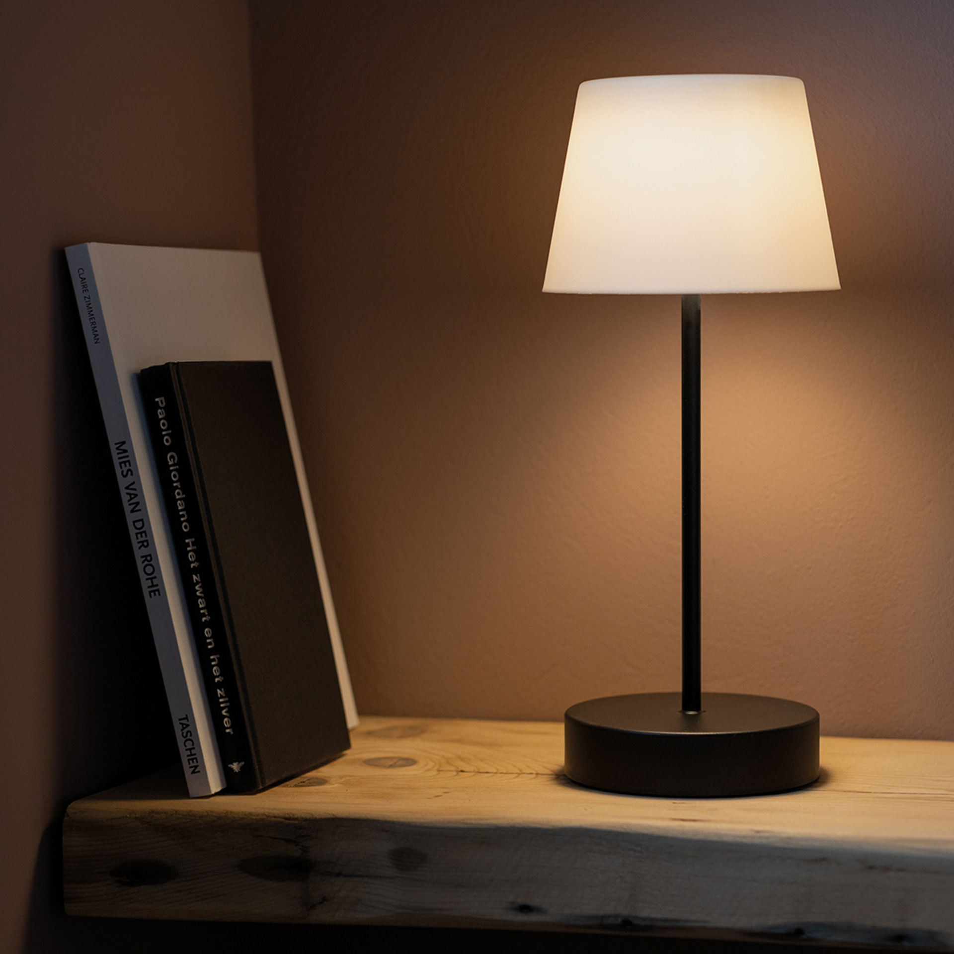 A 'pure' Oscar table lamp stands on a wooden board next to two books. The lamp has a black base and a white lampshade that emits a warm light.