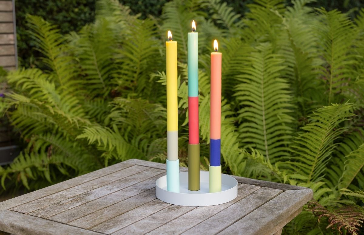 Drei leuchtende, mehrfarbige Kerzen stehen in einer runden, weißen Schale auf einem Gartentisch aus Holz. Links gelb, grau und hellblau, mittig türkis, rot und olivgrün, rechts orange, dunkelblau und hellgrün. Im Hintergrund üppige Farnblätter.