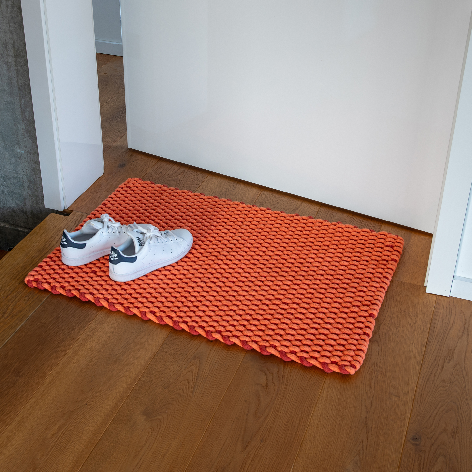 Un felpudo «Lida» de color coral descansa sobre un suelo de madera frente a una puerta blanca. Un par de zapatillas blancas descansan sobre el felpudo. El felpudo tiene una estructura tejida.