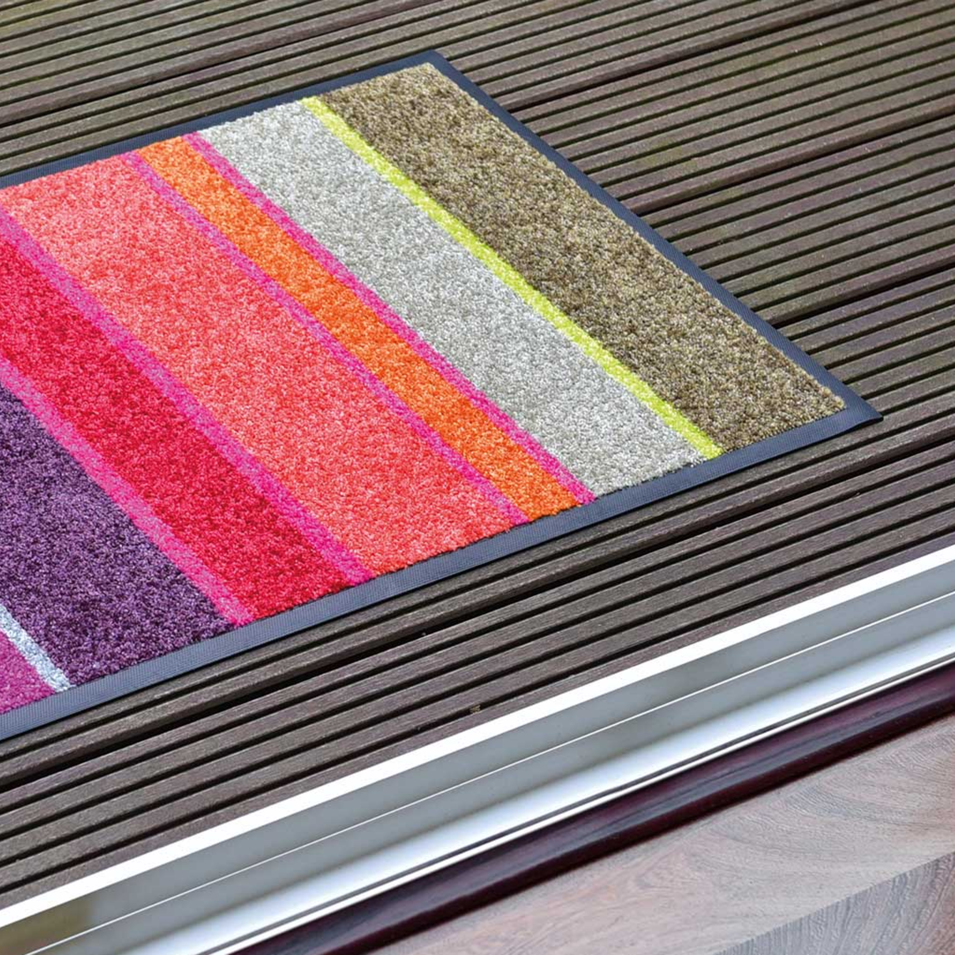 Eine 'Largo' Fußmatte mit bunten Streifen (lila, rot, orange, grau, braun) liegt auf einem Holzboden vor einer Türschwelle.