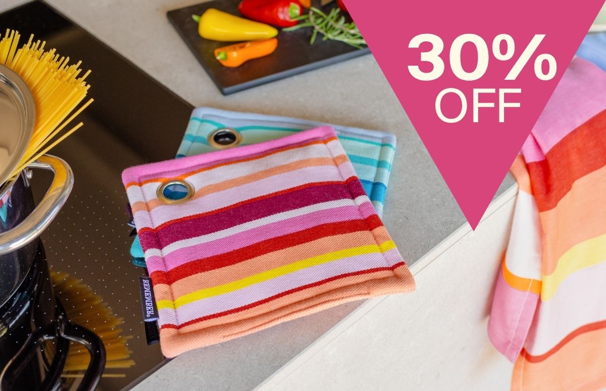 Zwei gestreifte Topflappen in Pink- und Blautönen mit Metallösen auf heller Küchenarbeitsplatte. Links ein Topf mit Spaghetti auf schwarzem Induktionskochfeld. Hinten Paprika und Kräuter. Oben rechts „30% OFF“.