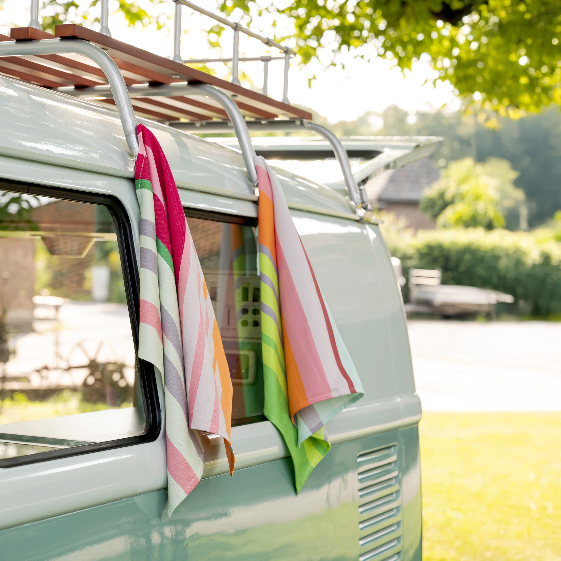 Deux lingettes sèches rayées sont accrochées à un bus VW vert menthe avec une galerie de toit. Les chiffons sont colorés avec des rayures roses, vertes, orange et grises. En arrière-plan, une prairie verte.