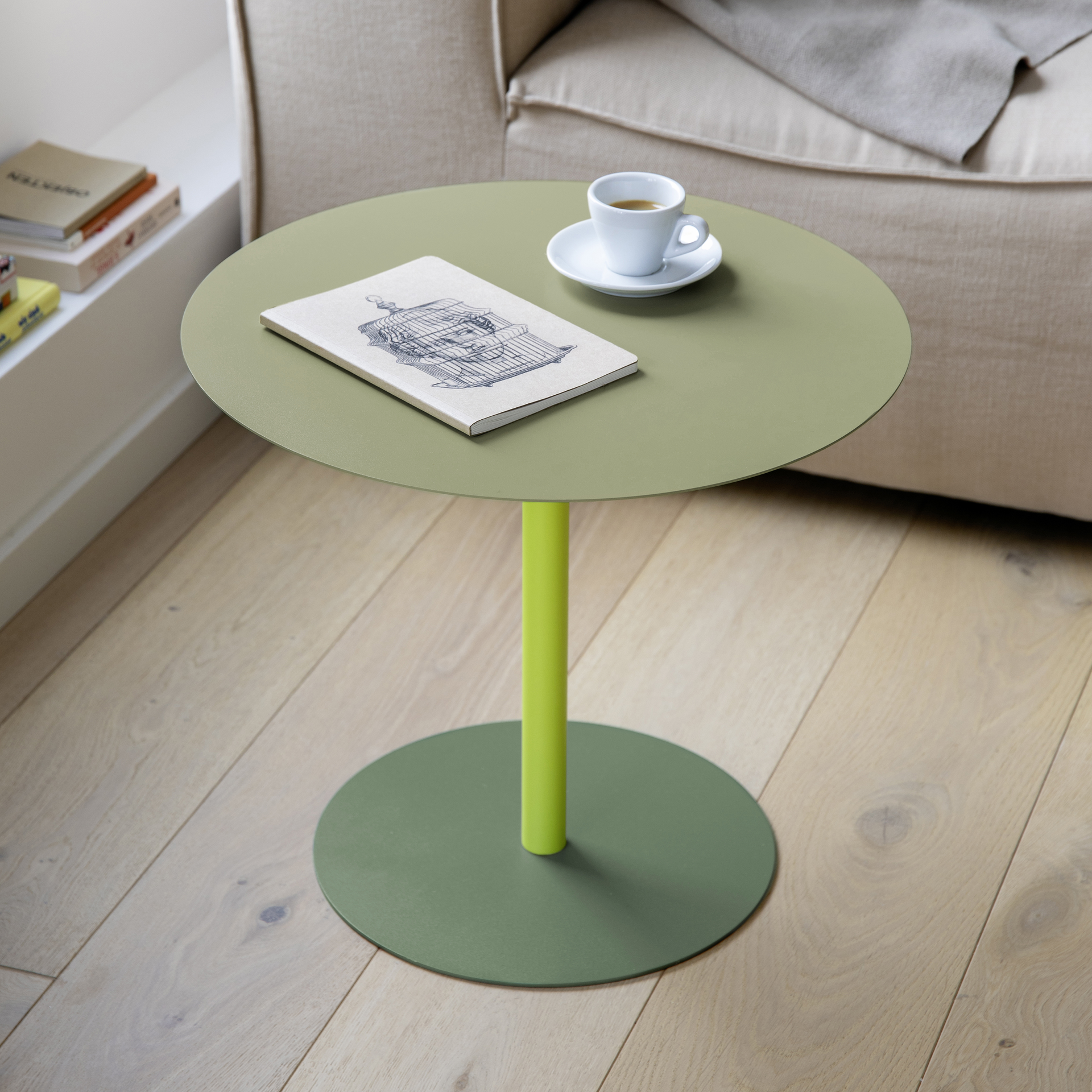 Ronde bijzettafel Rondo: mat olijfgroen tafelblad, limoengroene kolom, olijfgroene voet. Daarop een wit espressokopje en een notitieboekje. Op de achtergrond een beige fauteuil, een boekenkast en een lichte houten vloer.
