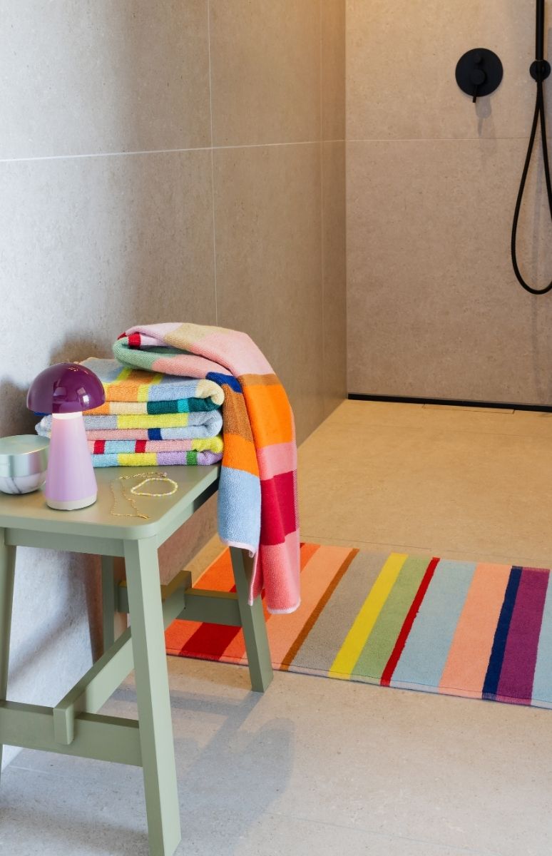 Modernes Badezimmer mit hellbraunen Fliesen. Links ein hellgrüner Hocker mit bunten, gestreiften Handtüchern, ein großes hängt herab. Darauf violette Pilzlampe, goldene Halskette und kleine Schale. Gestreifter Badteppich zur Dusche.