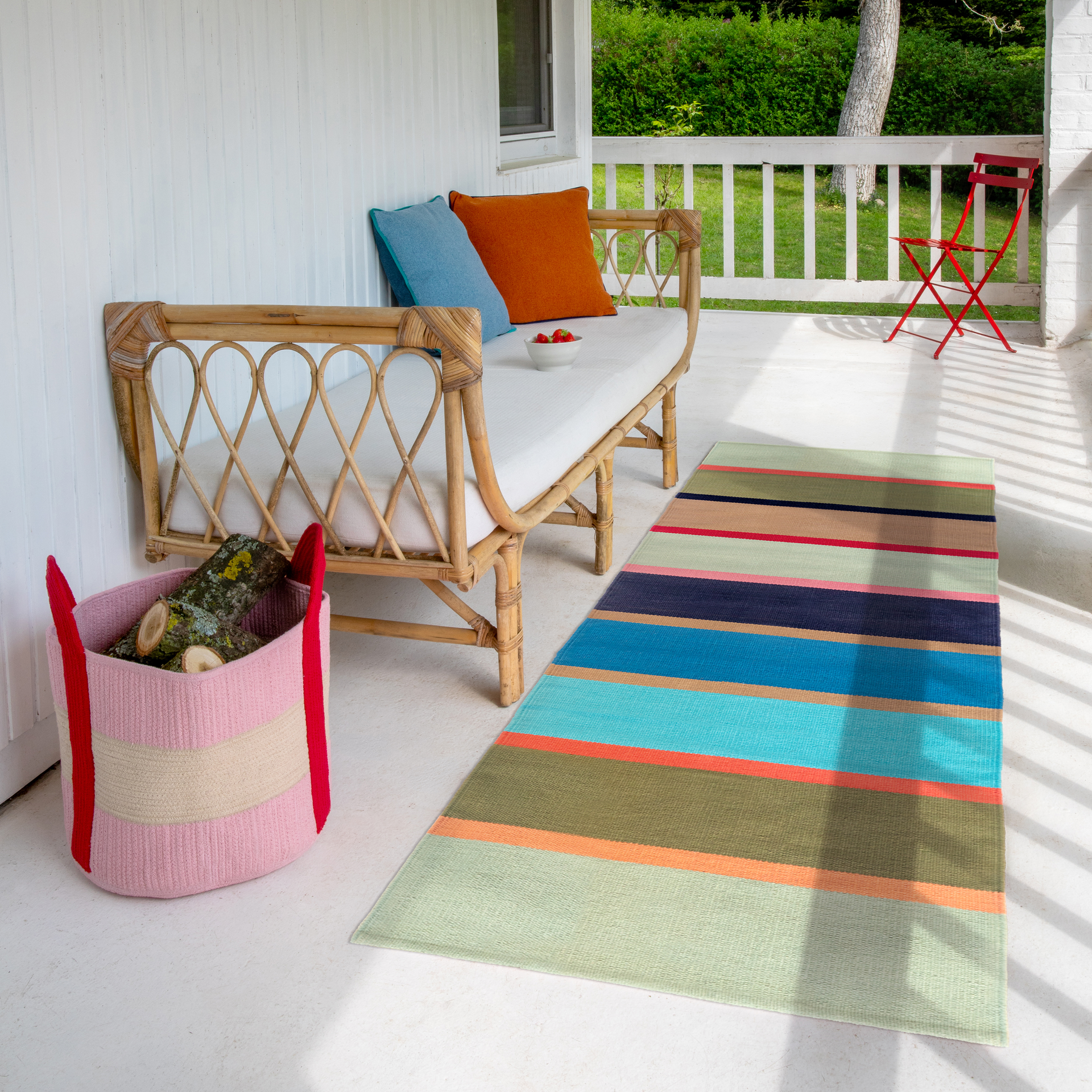 Véranda avec canapé en bambou, coussins blancs et coussins colorés ; corbeille de fraises. À gauche, panier de bûches. Tapis à rayures colorées, rayons de soleil, chaise pliante rouge à l'arrière.