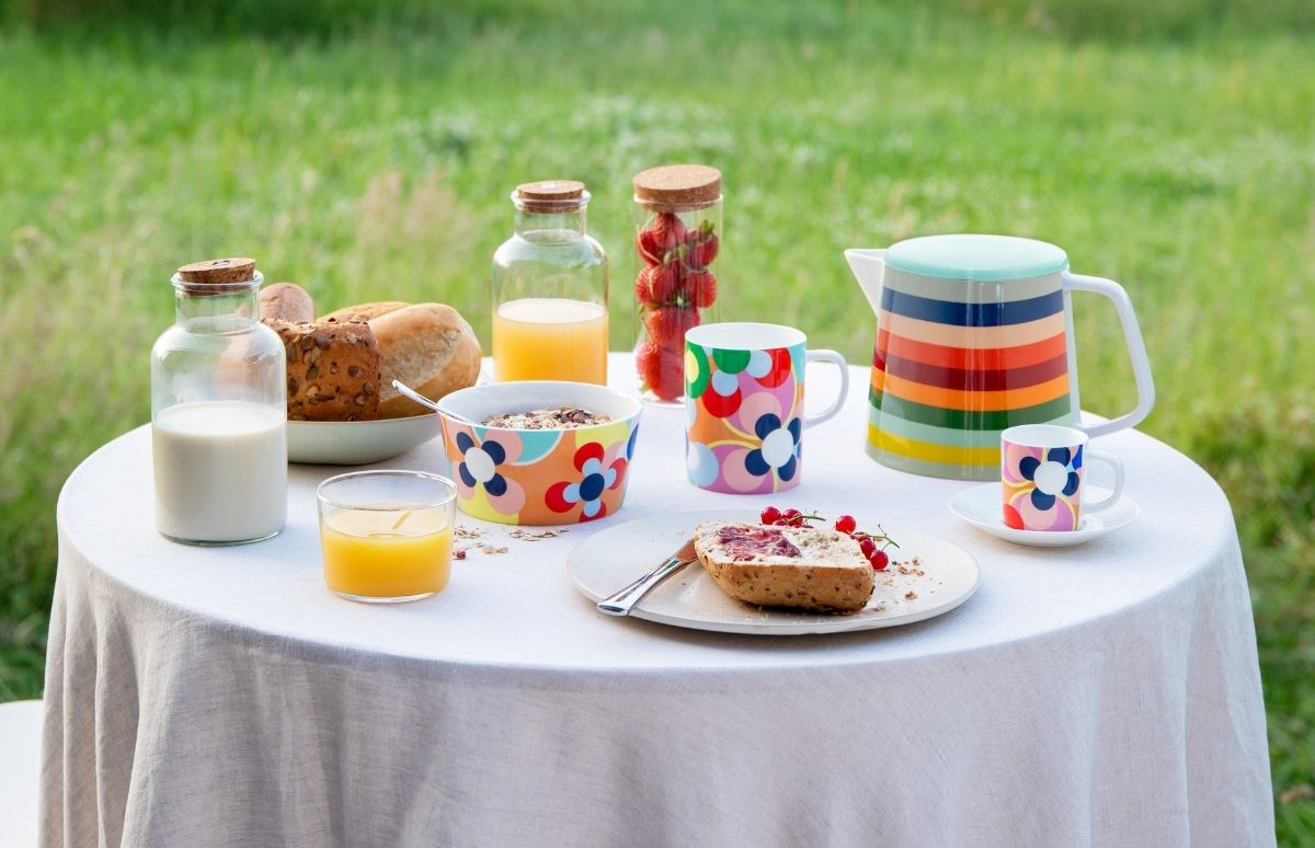 Gedeckter Frühstückstisch im Freien auf grüner Wiese. Auf weißem Tischtuch stehen farbenfrohes Geschirr mit Blumenmuster und eine gestreifte Kanne. Daneben: Milch, Orangensaft, Müsli, Brot, Erdbeeren und Marmeladenbrot mit Johannisbeeren.