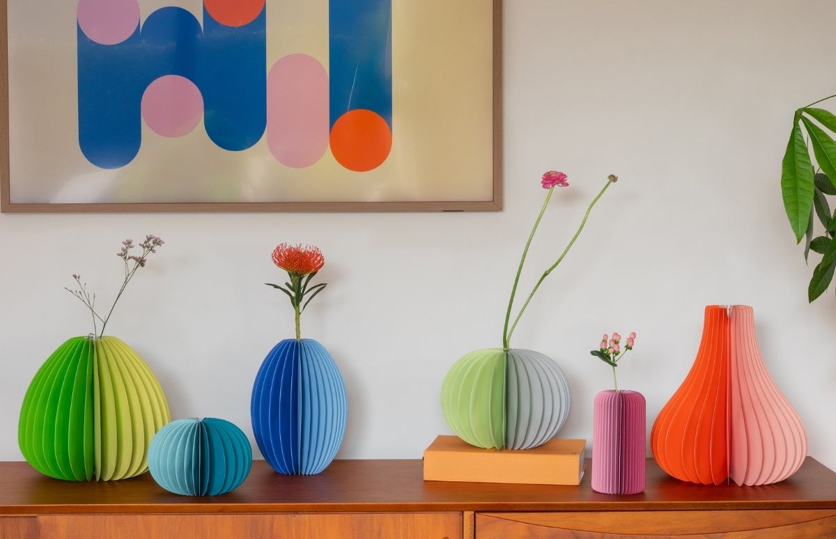 Gefaltete Papier-Vasen in leuchtenden Farben wie Grün, Blau, Pink und Orange stehen auf einer Holzkommode. Einige enthalten einzelne Blumen oder getrocknete Zweige, darunter eine rote Protea und pinke Blüten. Darüber hängt ein abstraktes Bild.