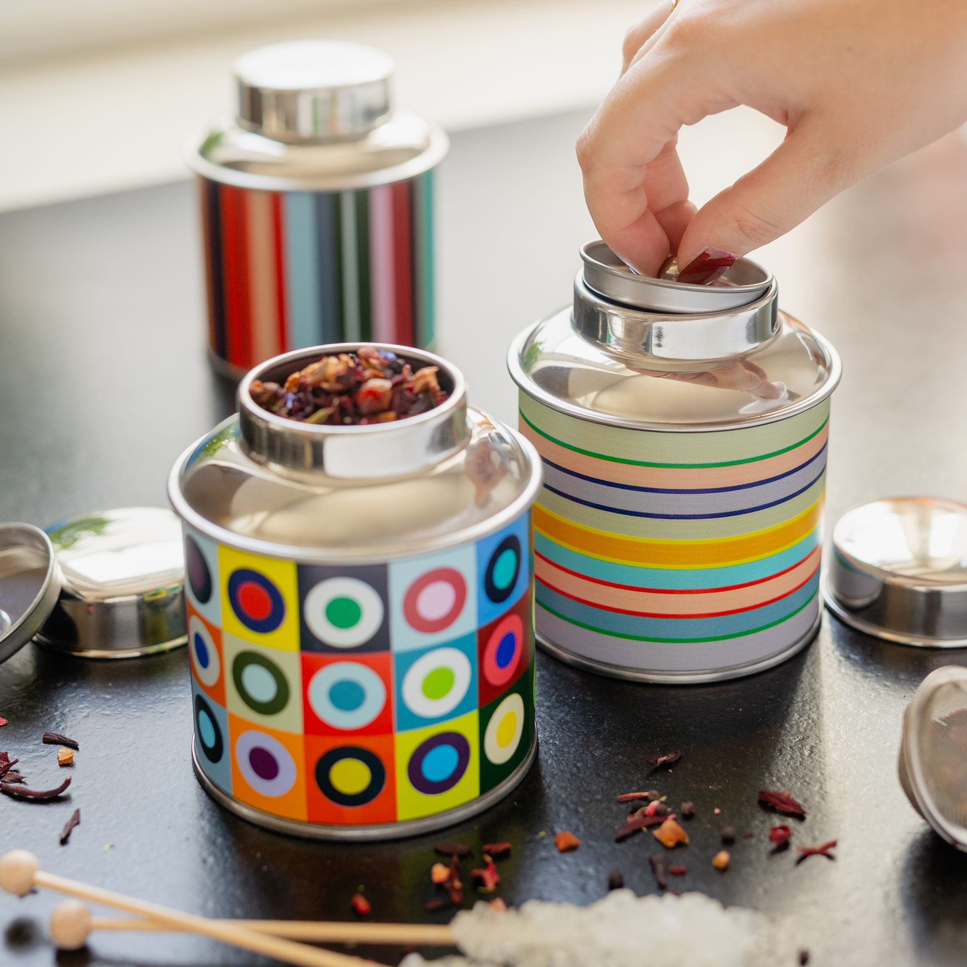 Trois boîtes à thé colorées « REMEMBER » sur fond sombre. Deux boîtes inox ouvertes à l'avant : l'une avec du thé aux fruits en vrac, l'autre remplie par une main. Motif : cercles et rayures. Thé et sucre éparpillés.