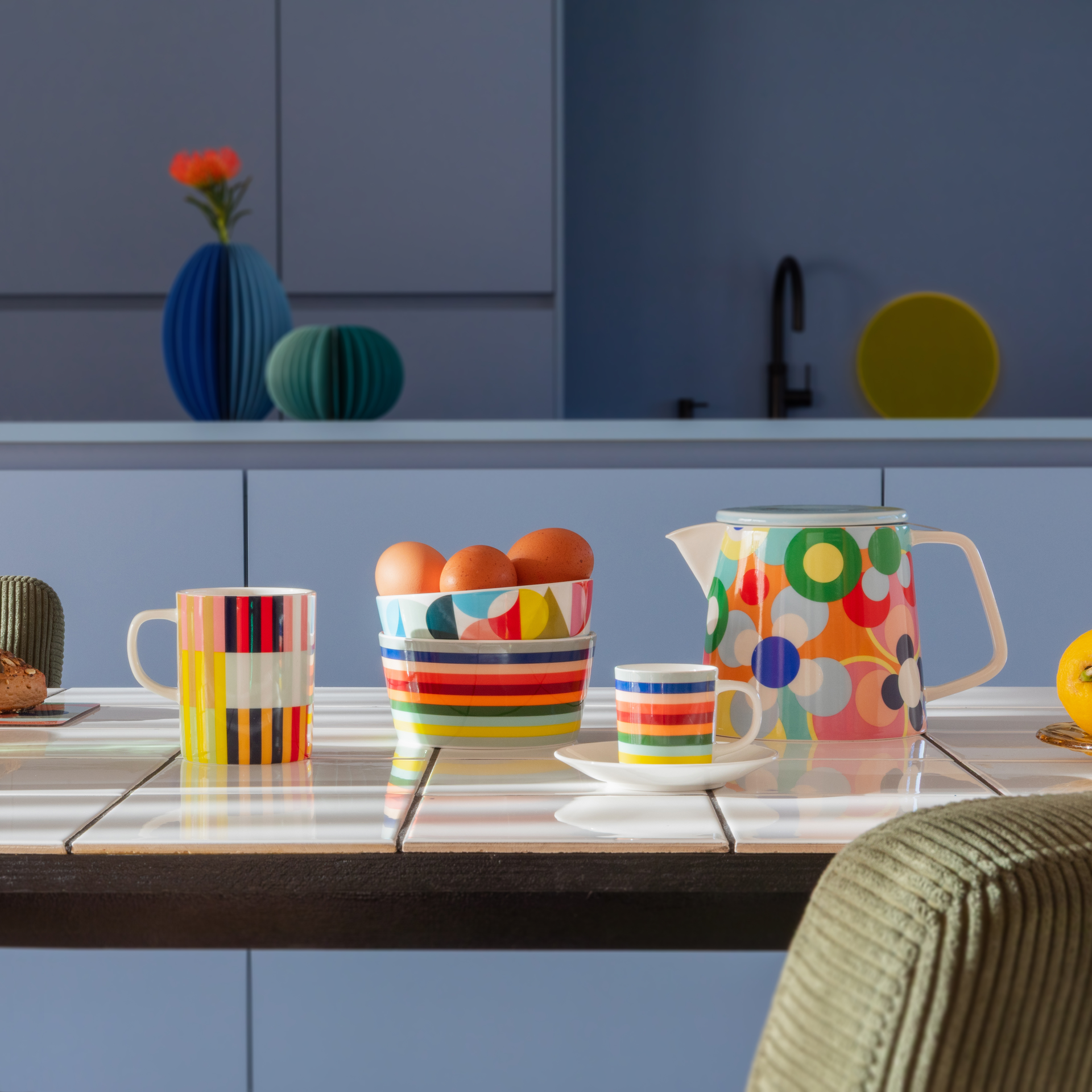 Tavolo da cucina con stoviglie. Una brocca con motivo circolare, tazze a righe, ciotole con uova. Sullo sfondo armadi e decorazioni blu.